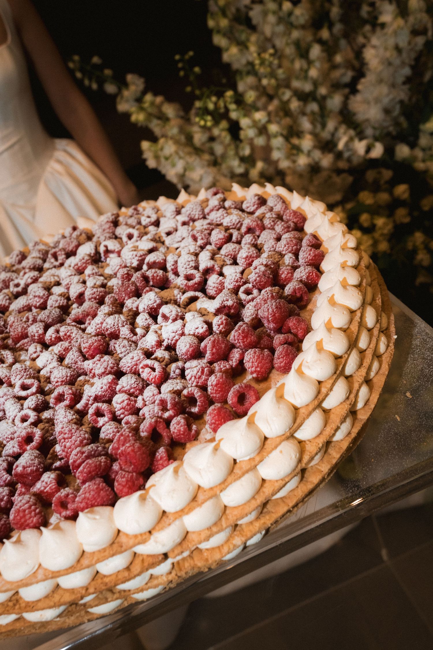Large round raspberry tart dusted with powdered sugar and topped with whipped cream dollops, displayed on a tray with white flowers behind.