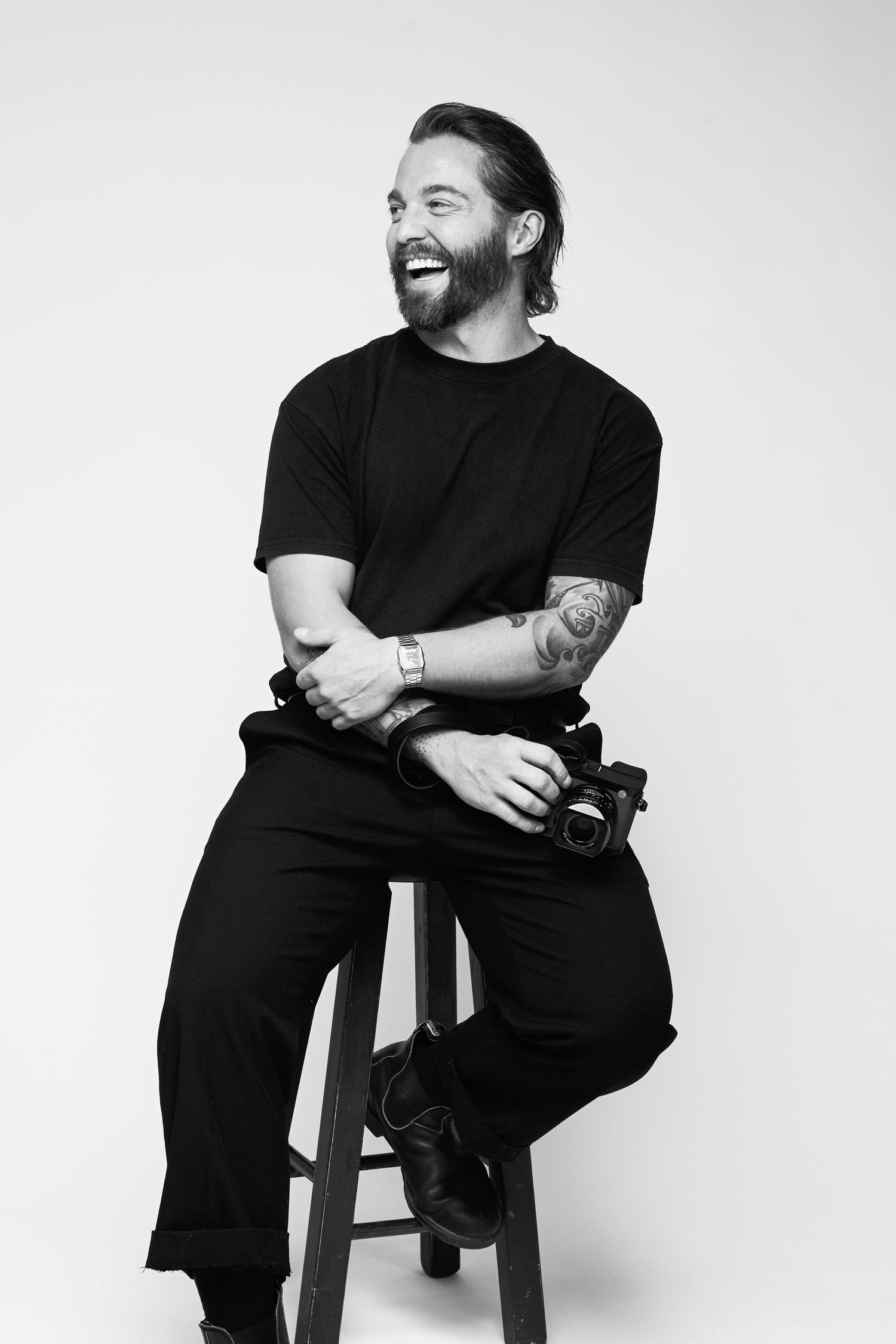 Black and white portrait of a bearded man with tattoos sitting on a stool, laughing, wearing all black, holding a camera.