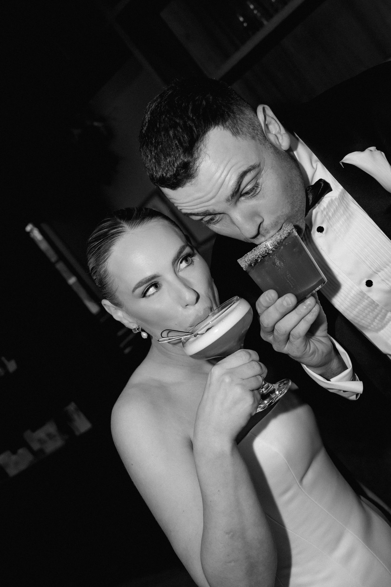 Black and white photo of a bride and groom sharing cocktails at their wedding reception in a romantic, dimly lit setting.