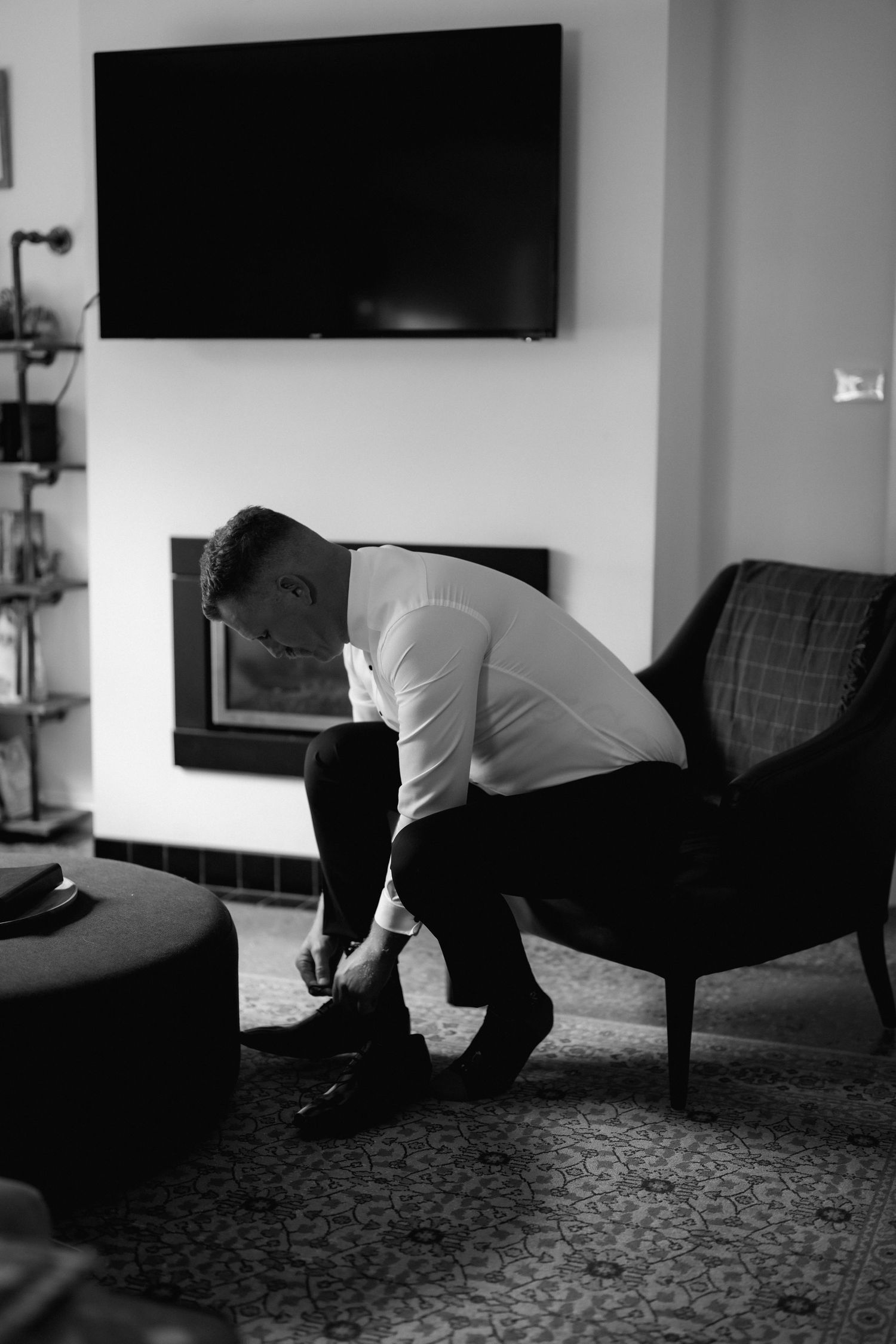 A person in a white shirt leans down to tie their shoes in a modern living room with a wall-mounted TV and armchair.