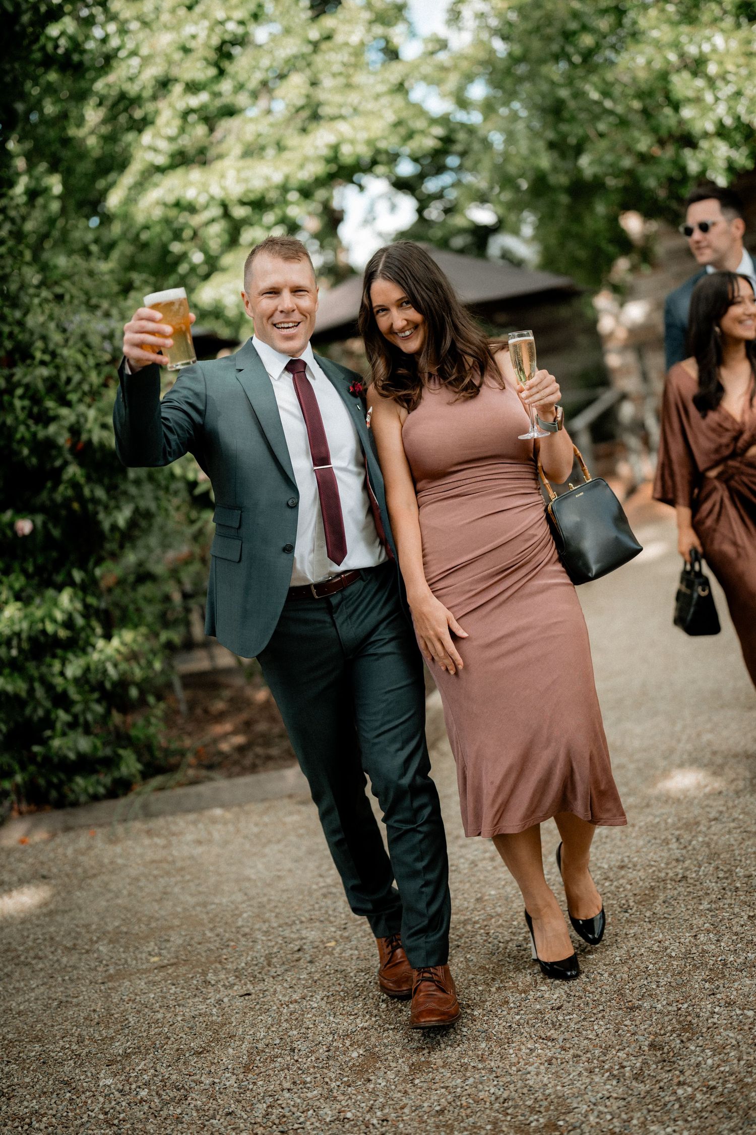 Wedding party in formal attire poses playfully on a garden path with groomsmen in suits and bridesmaids in brown dresses.