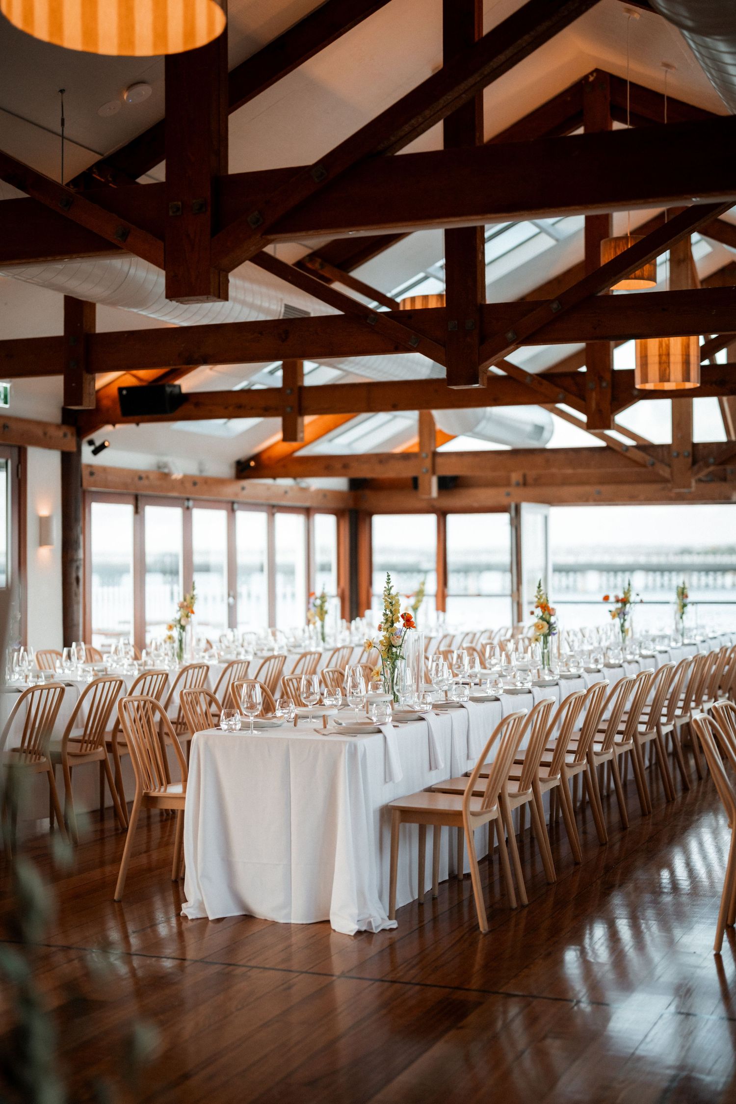 Large rustic reception venue with exposed wooden beams and rows of white-clothed tables.
