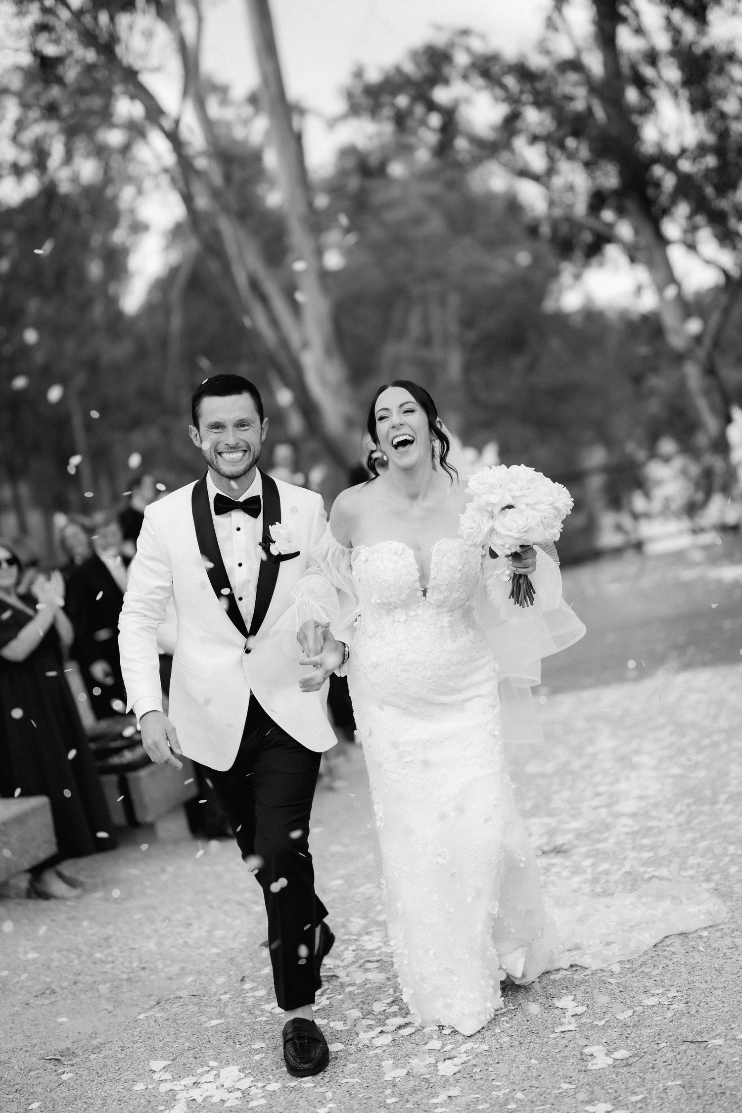 Black and white photo of a couple running through flower petals in formal attire.