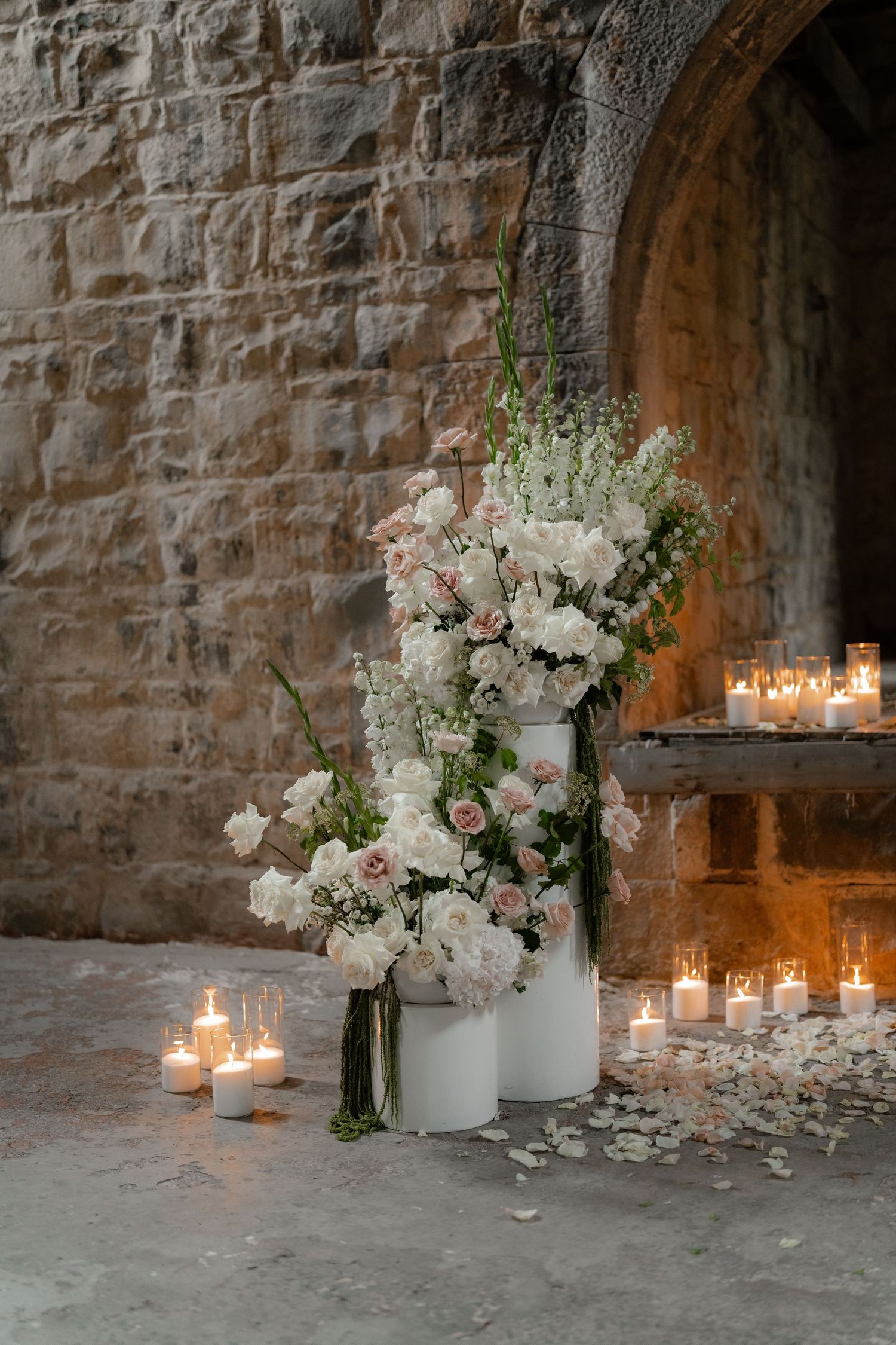 Elegant floral arrangement with white and pink flowers in a vase alongside glowing candles against a stone wall.