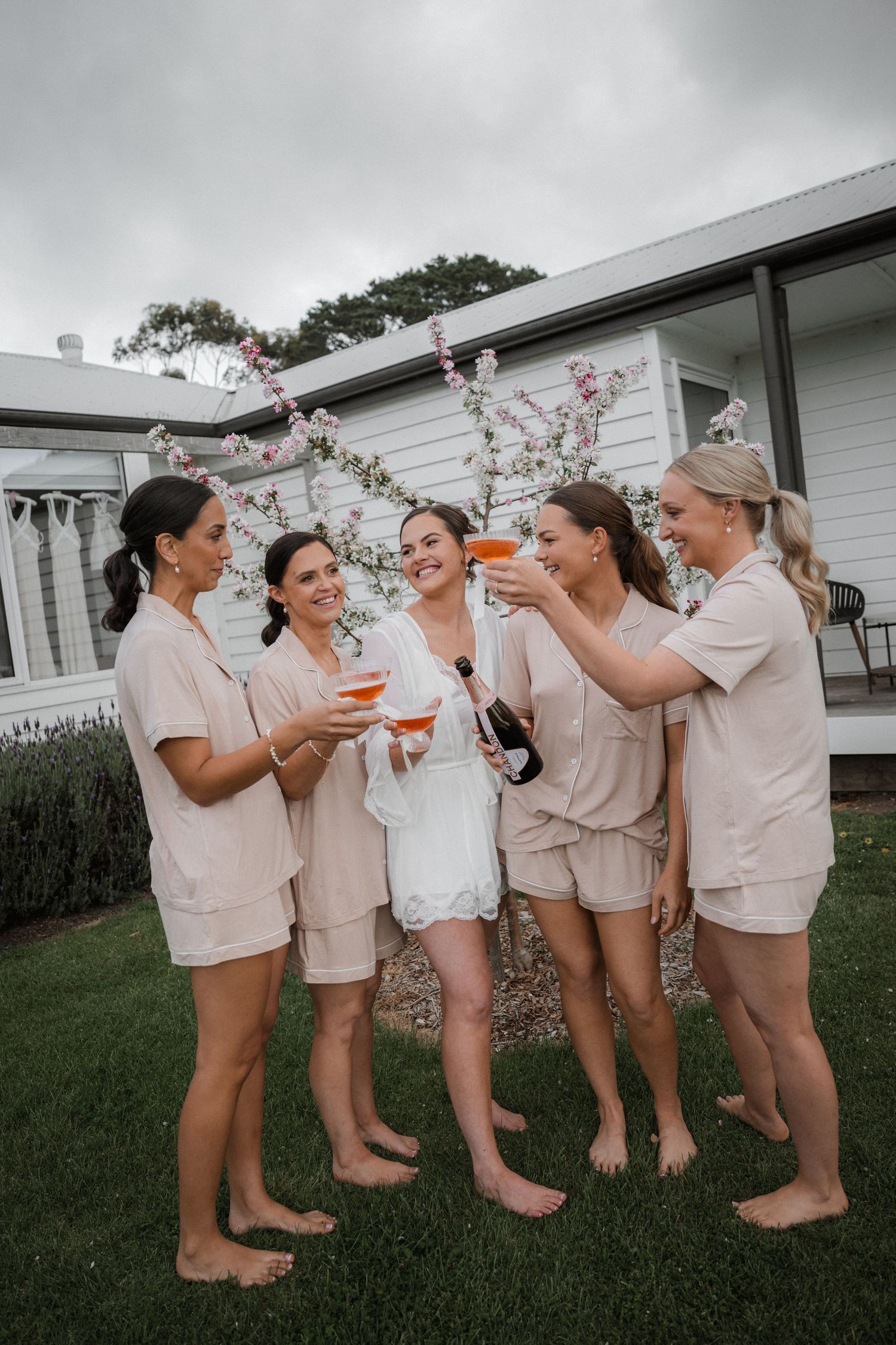 Bridesmaids in beige outfits laugh and celebrate together on a grassy lawn.