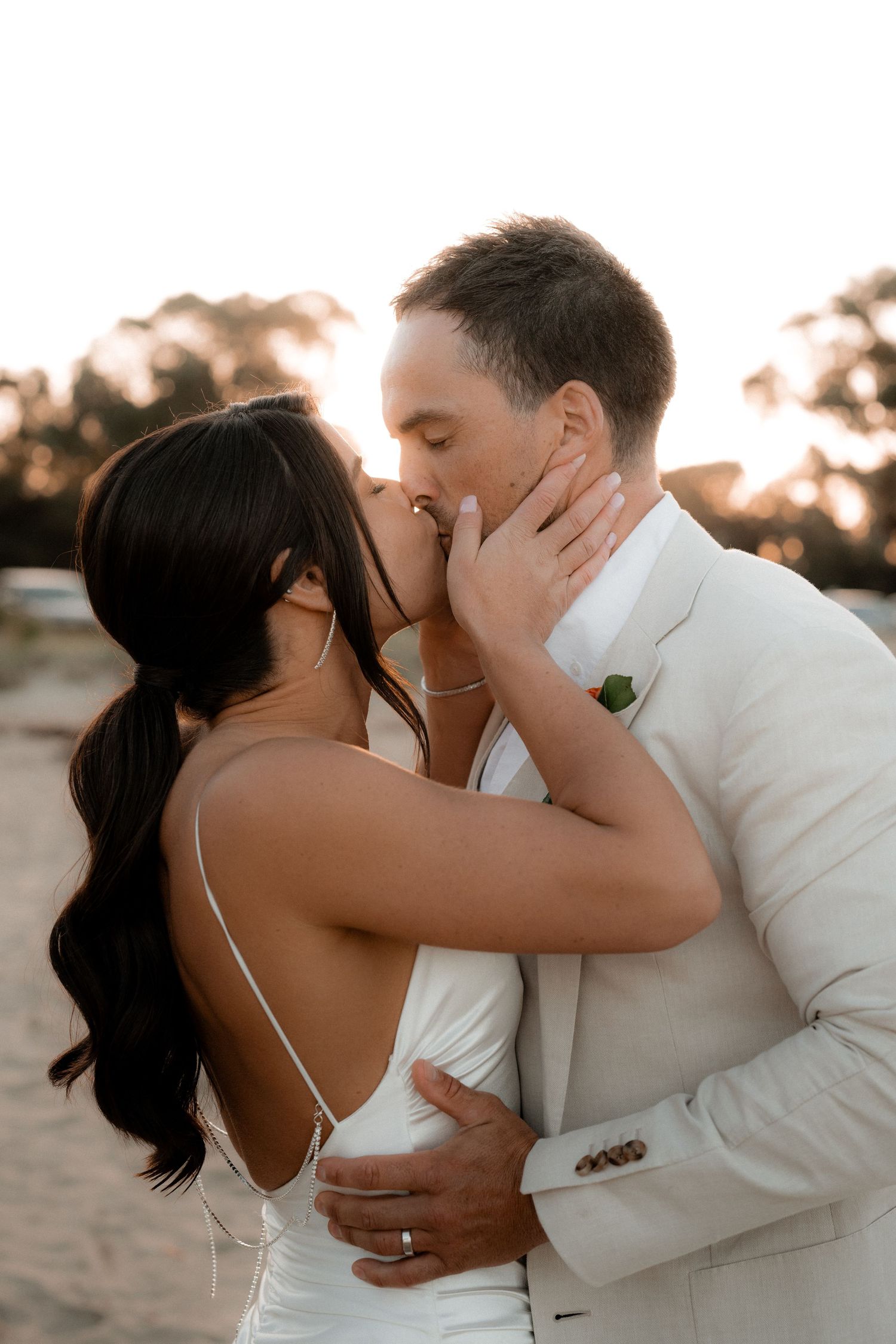 Romantic sunset beach portraits of newlyweds embracing and kissing in formal wedding attire.