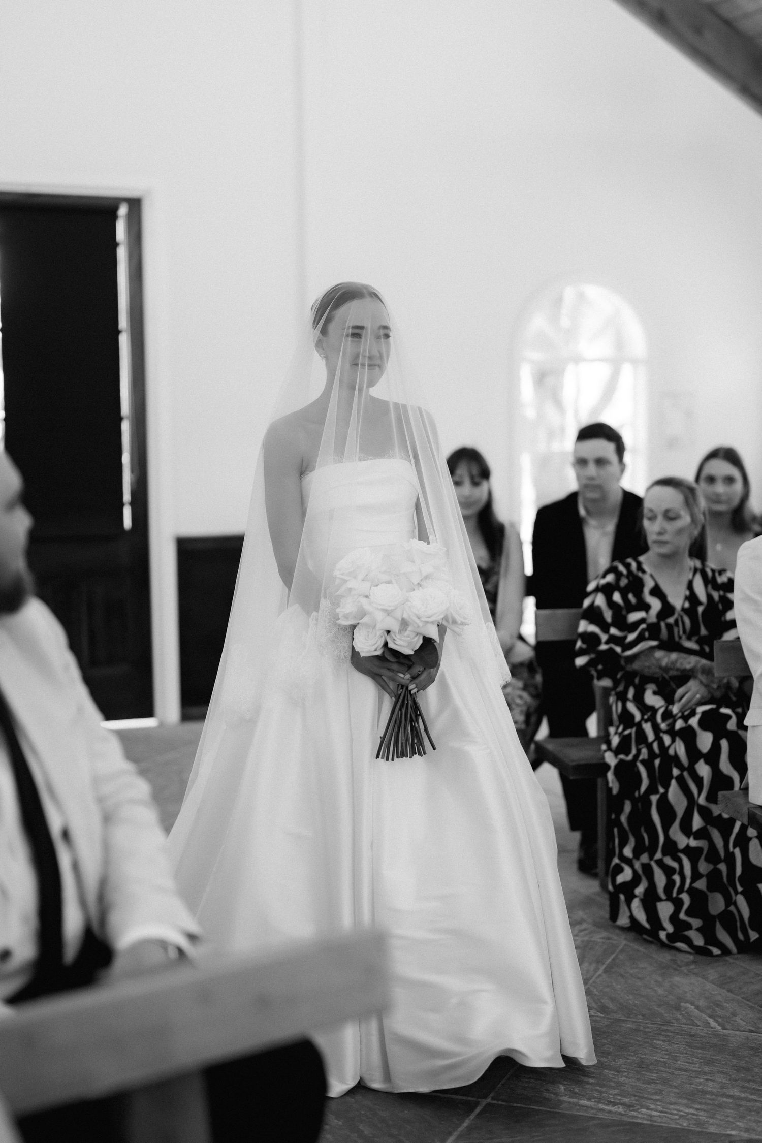 A bride in a classic white gown stands with her wedding party in a bright indoor setting.