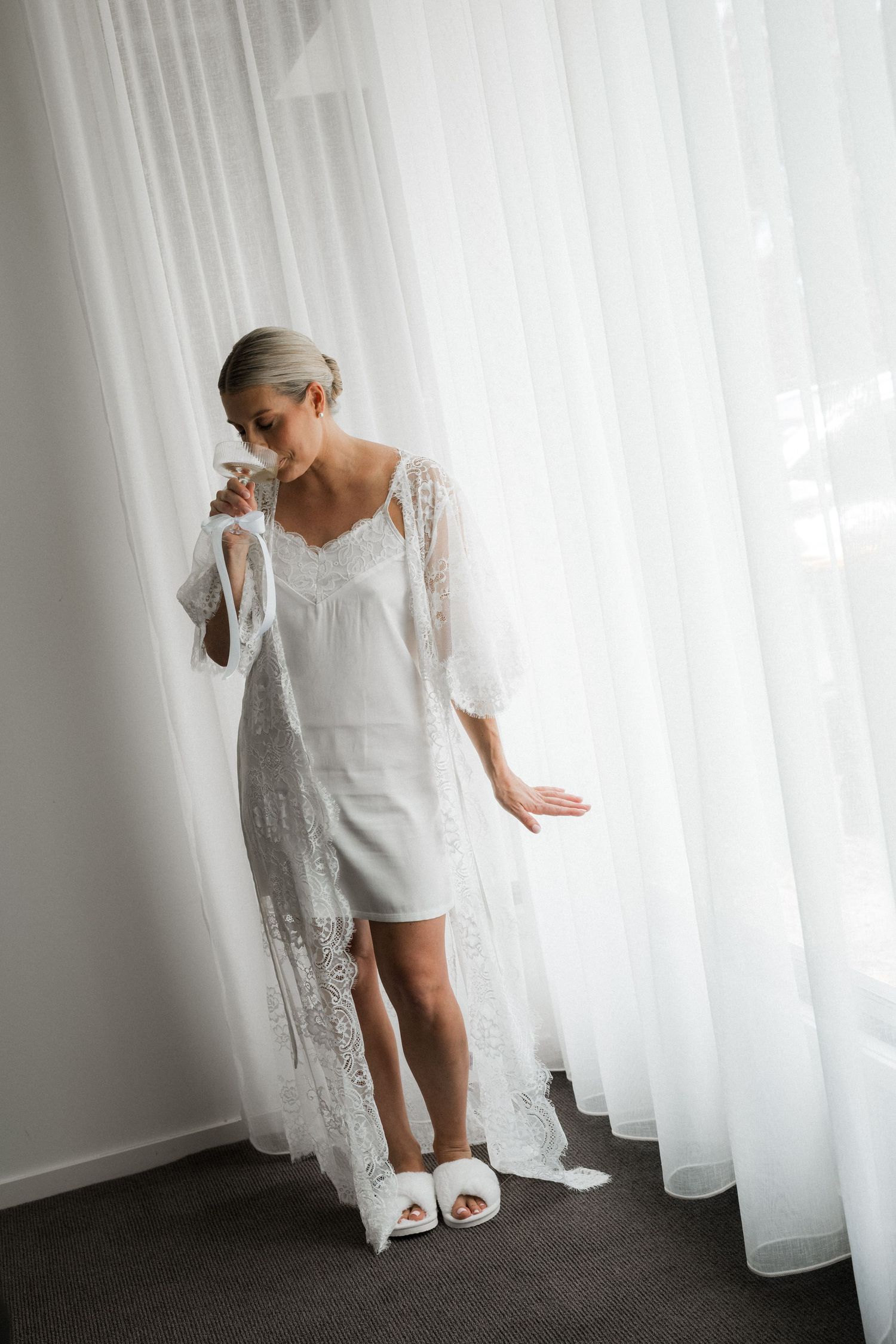 Elegant white lace wedding dress displayed against sheer curtains in natural light.