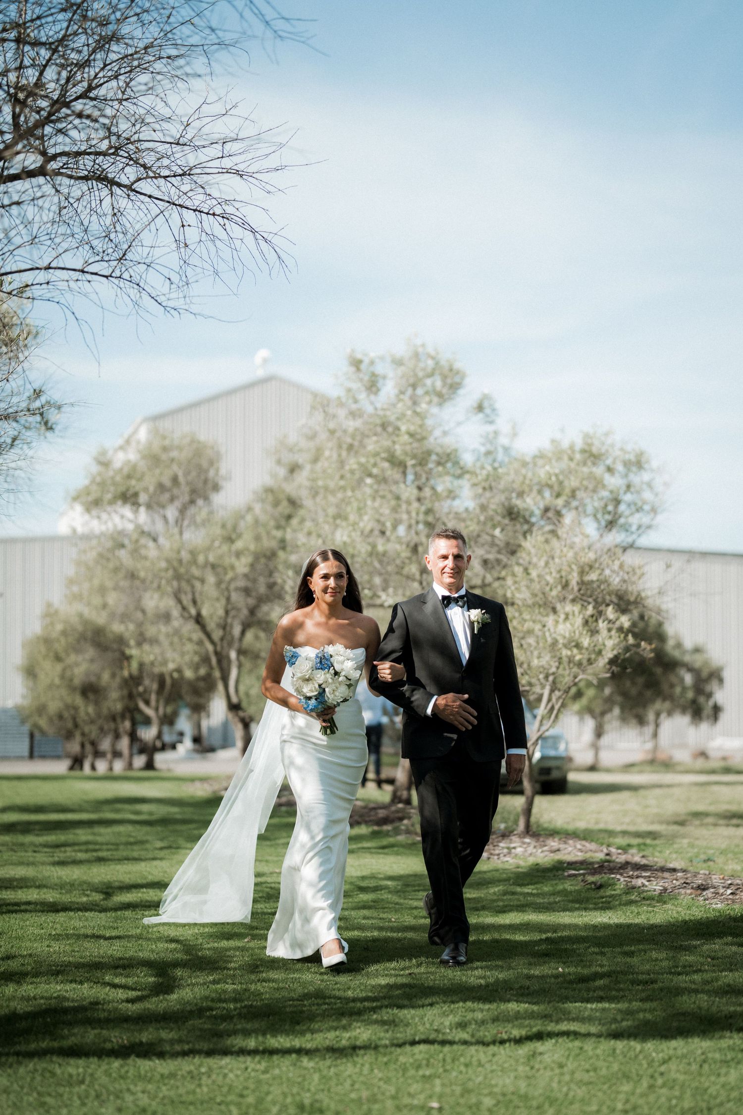 A couple walks together on a green lawn with trees and buildings in the background.