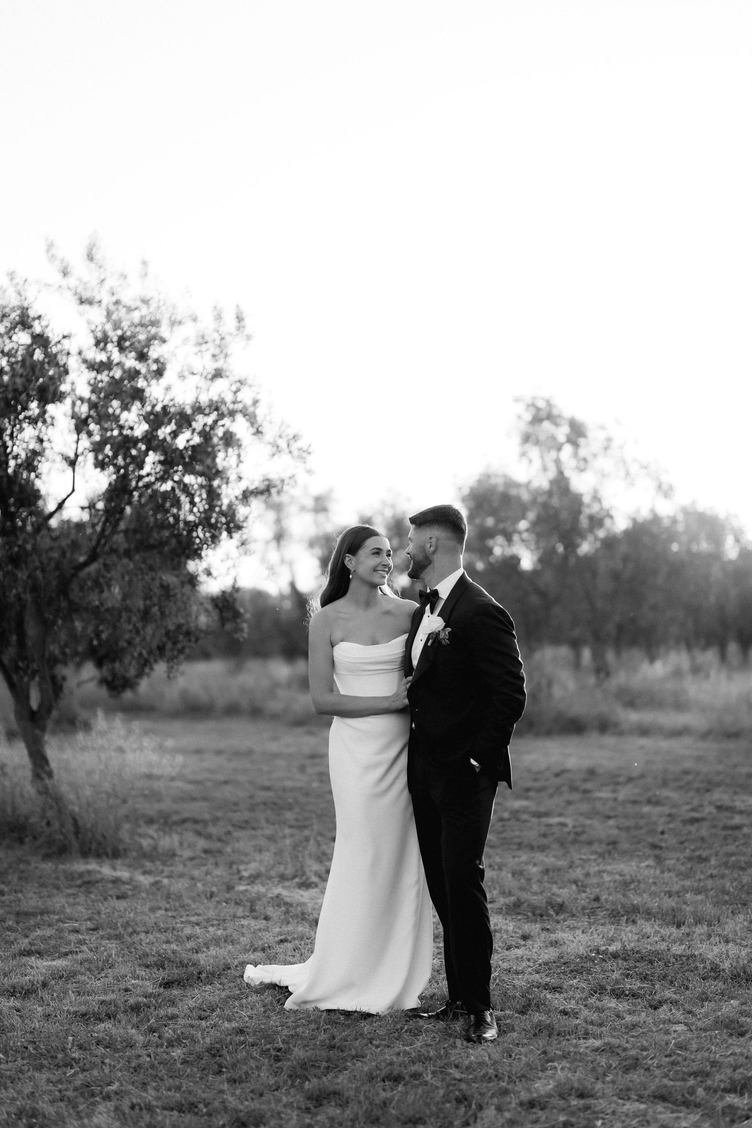 Black and white portrait of a couple in wedding attire walking through a field.