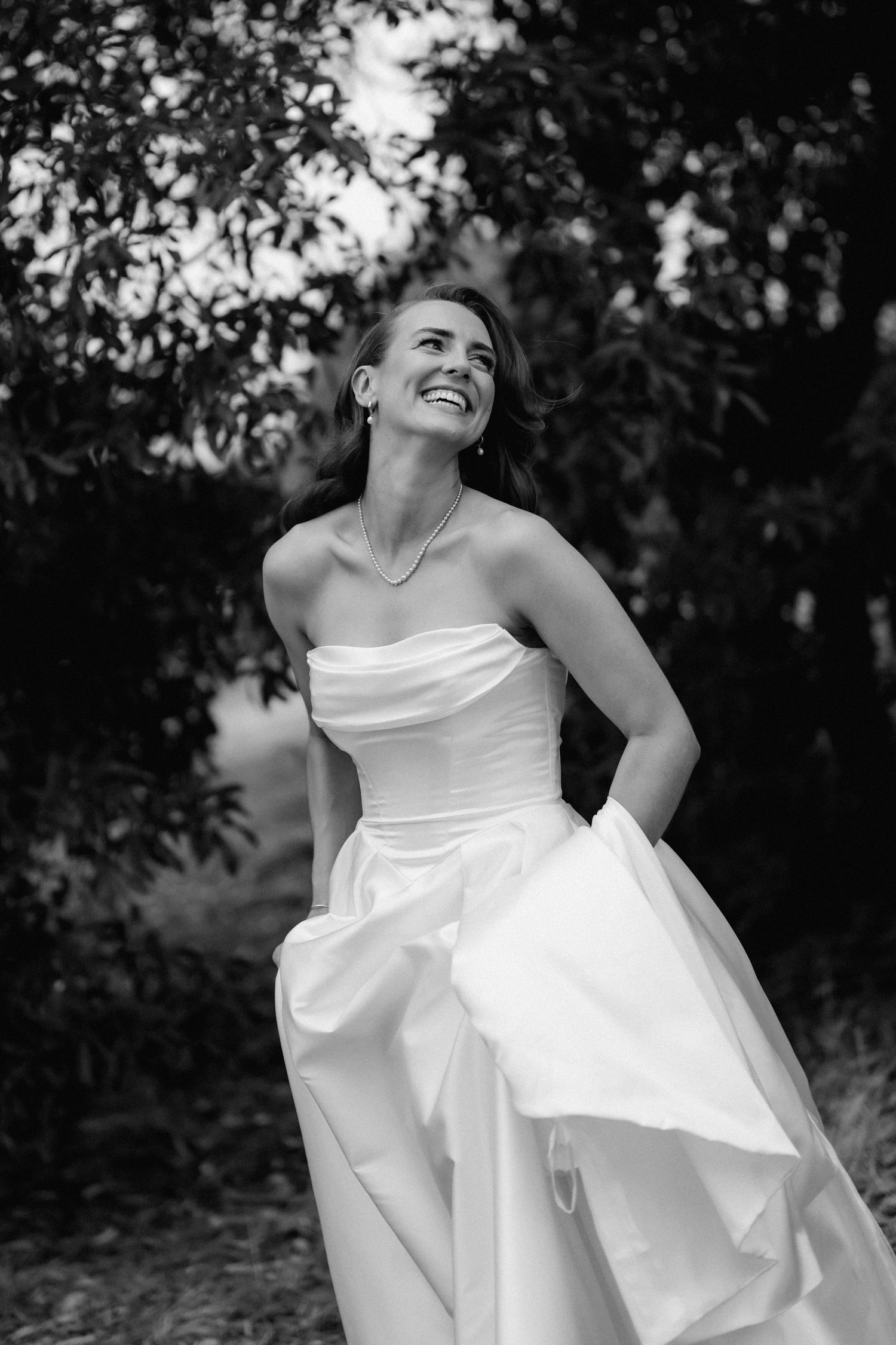 Black and white photo of a joyful bride in a strapless ball gown laughing outdoors among lush green trees.