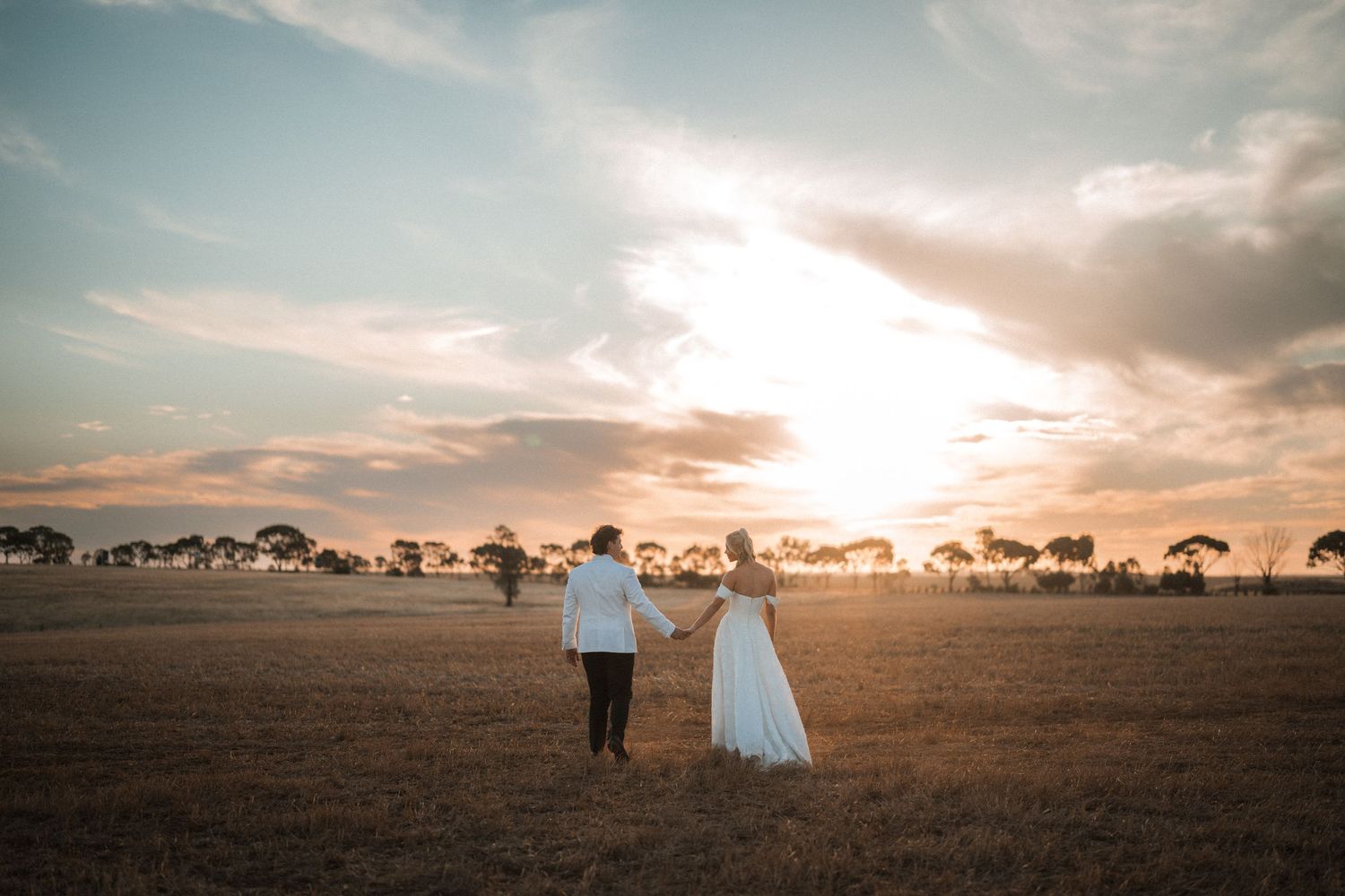 Bride and groom holding hands walking through golden field at sunset.