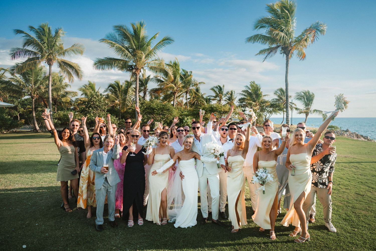 Large wedding party celebrating on a tropical lawn with palm trees and ocean backdrop.