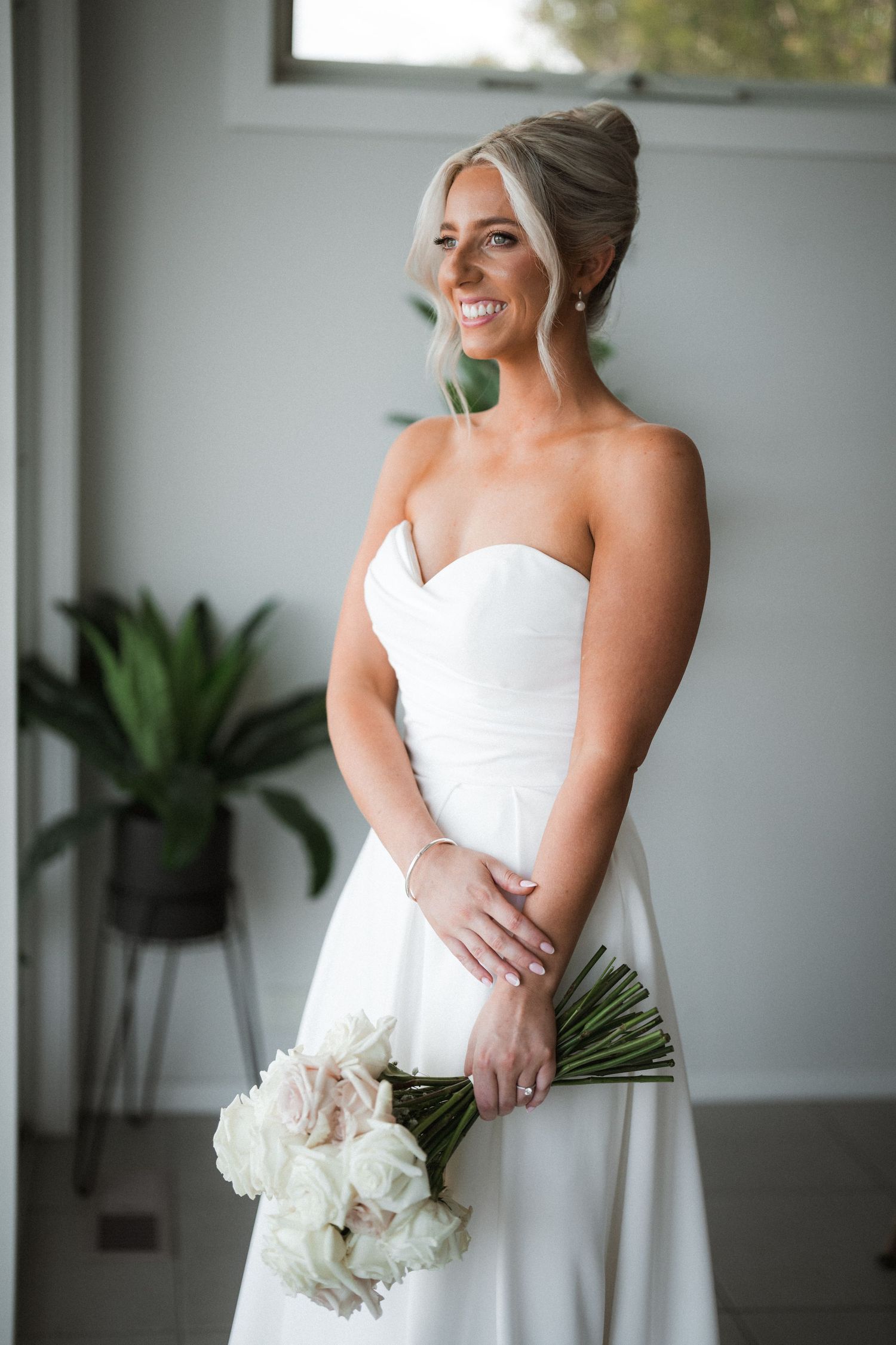 Elegant bride in a strapless white wedding gown smiles while holding a bouquet of white peonies near a window.