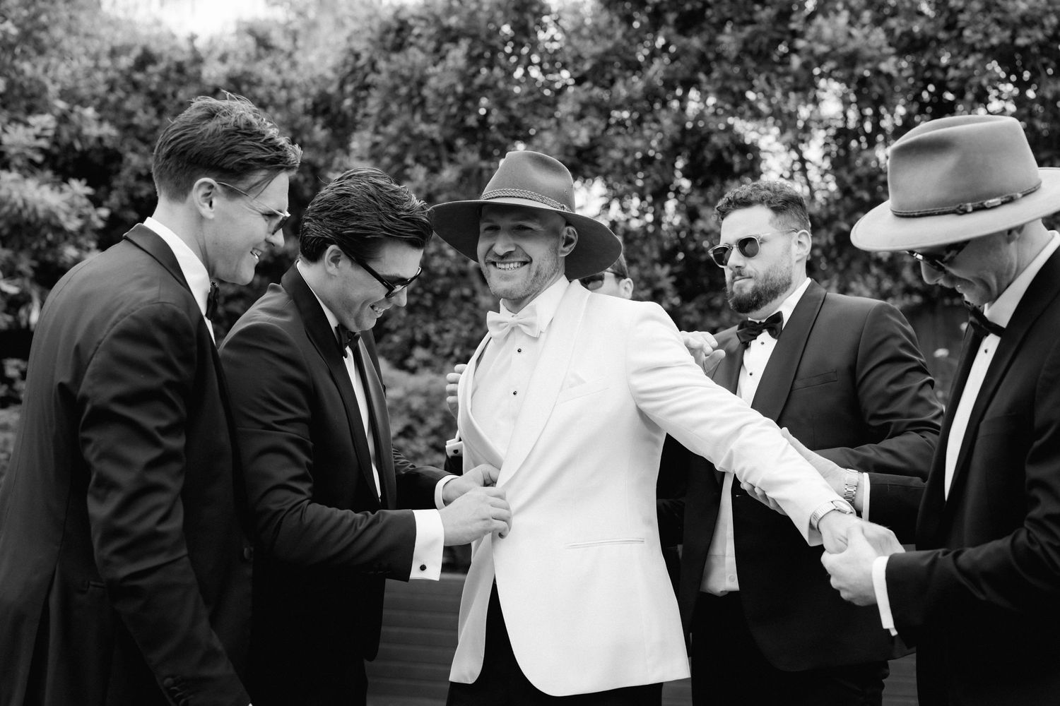 A candid black and white photo shows a group of five men in formal attire laughing and interacting at an outdoor gathering.