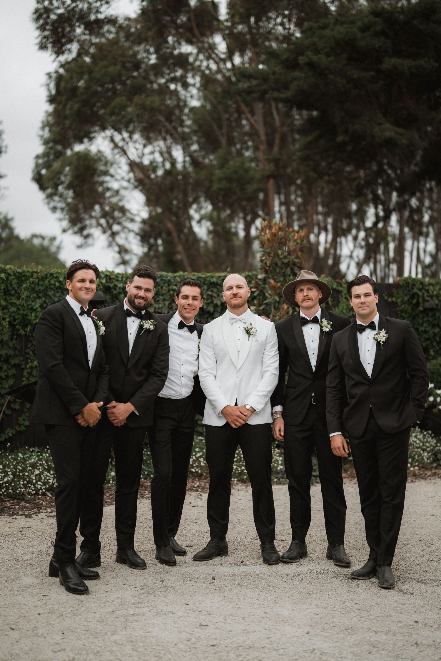 A wedding party of groomsmen in formal black tuxedos and one in white standing together outdoors with trees in background.