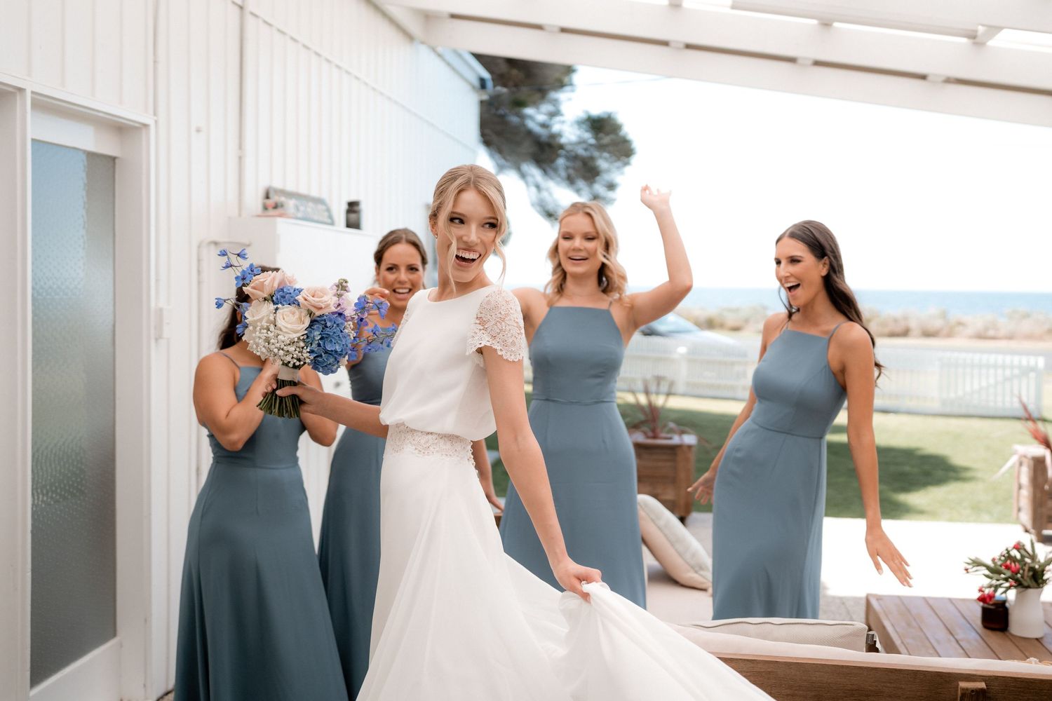 Wedding party in dusty blue bridesmaid dresses celebrating on a sunny outdoor patio.