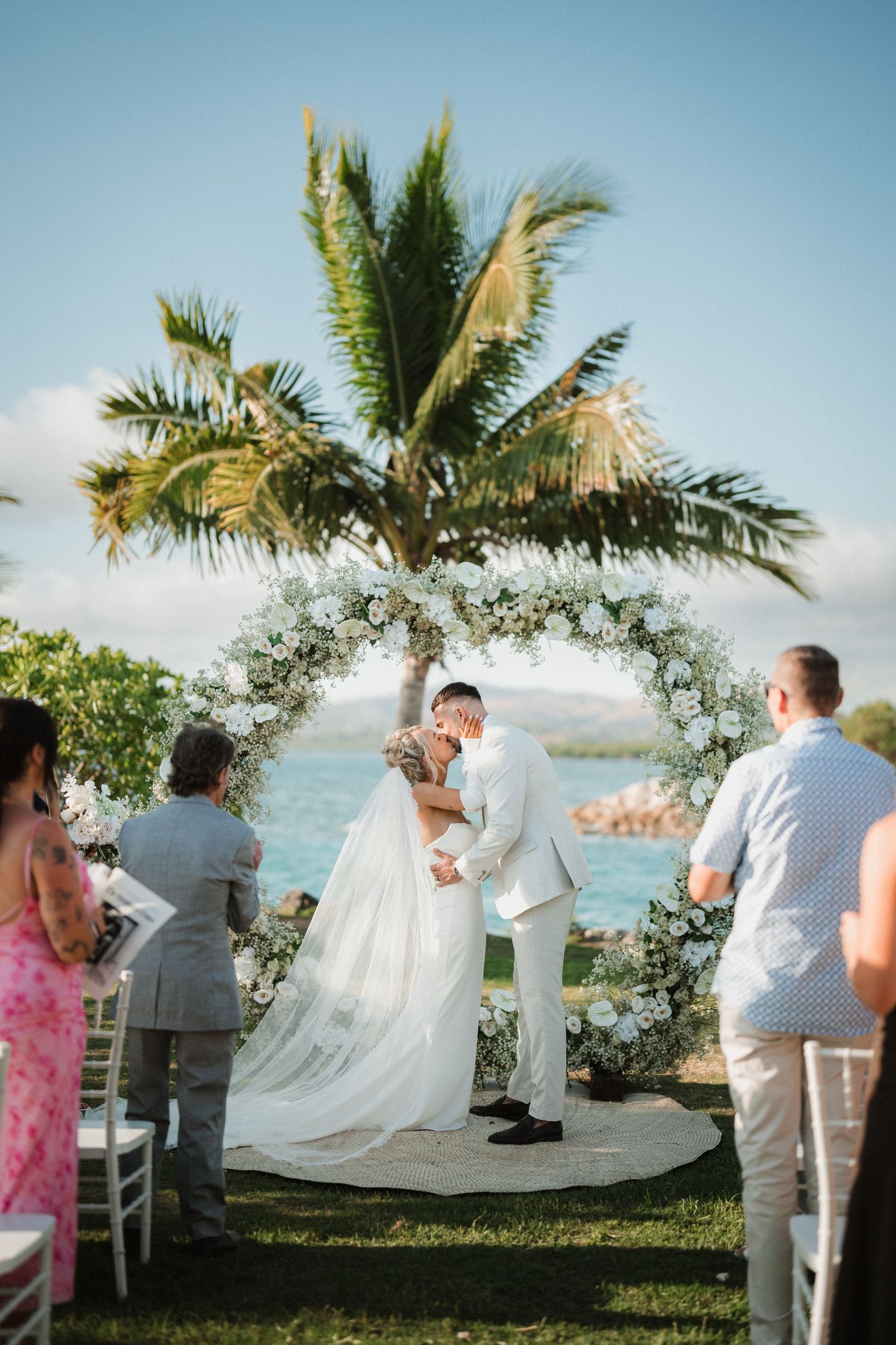 A beautiful outdoor beach wedding ceremony under a floral arch with palm trees and ocean views in Hawaii.