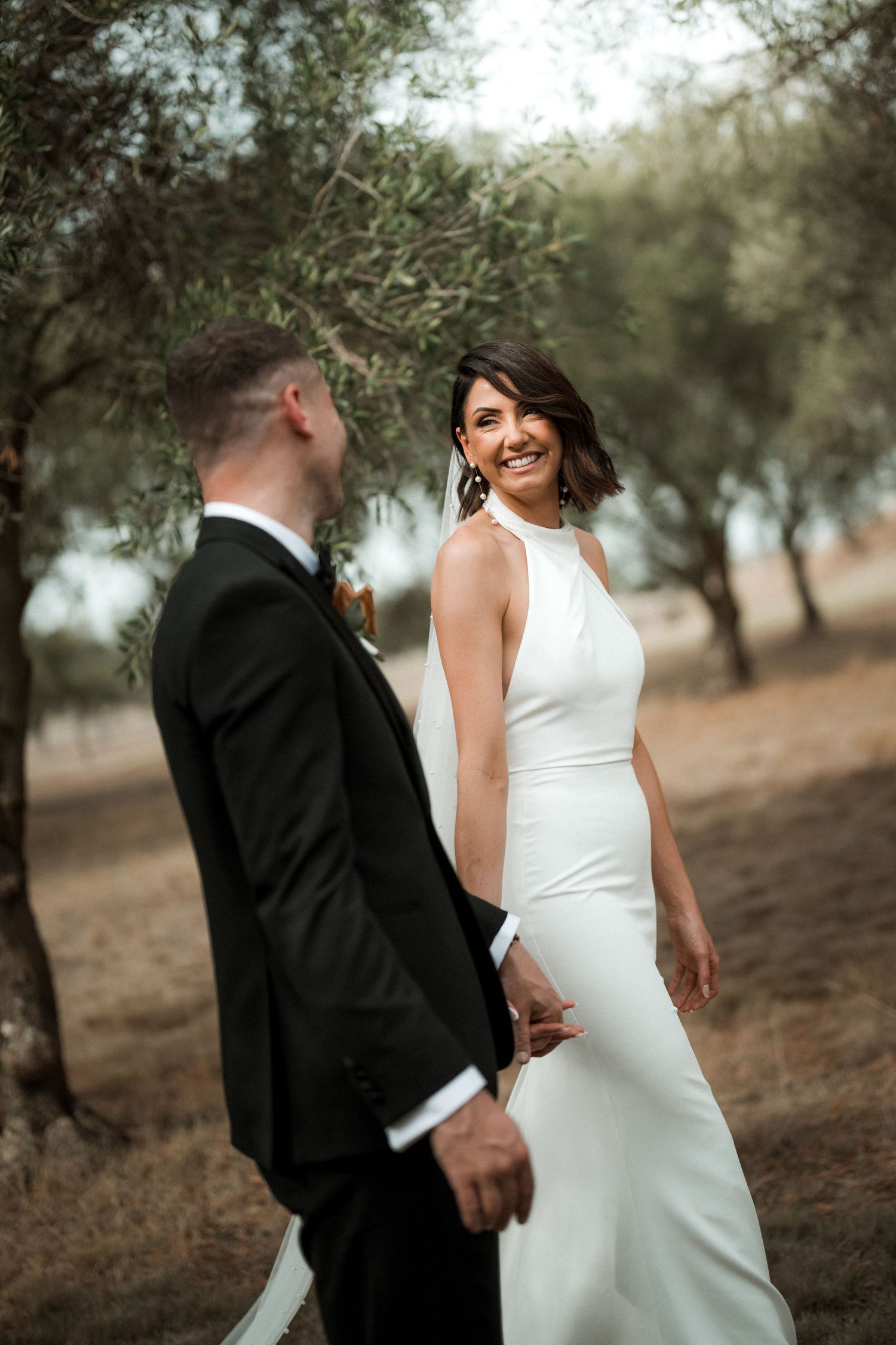 Elegant couple in wedding attire pose together in an outdoor setting with olive trees in the background.
