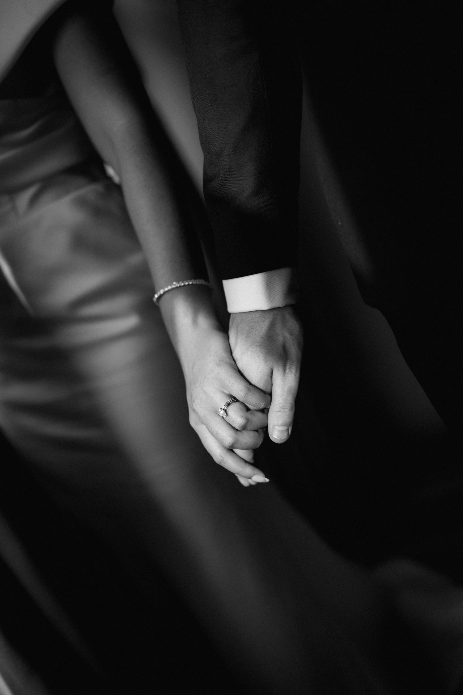 Black and white close-up of clasped hands wearing a delicate bracelet and ring against dark fabric.