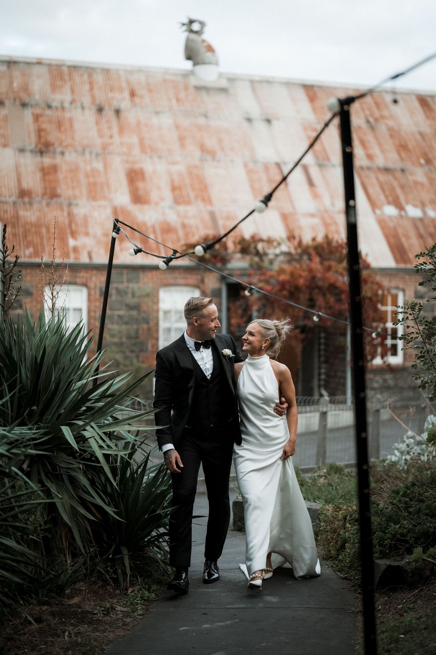 A newlywed couple walks down a garden path together under string lights with a rustic brick building backdrop.