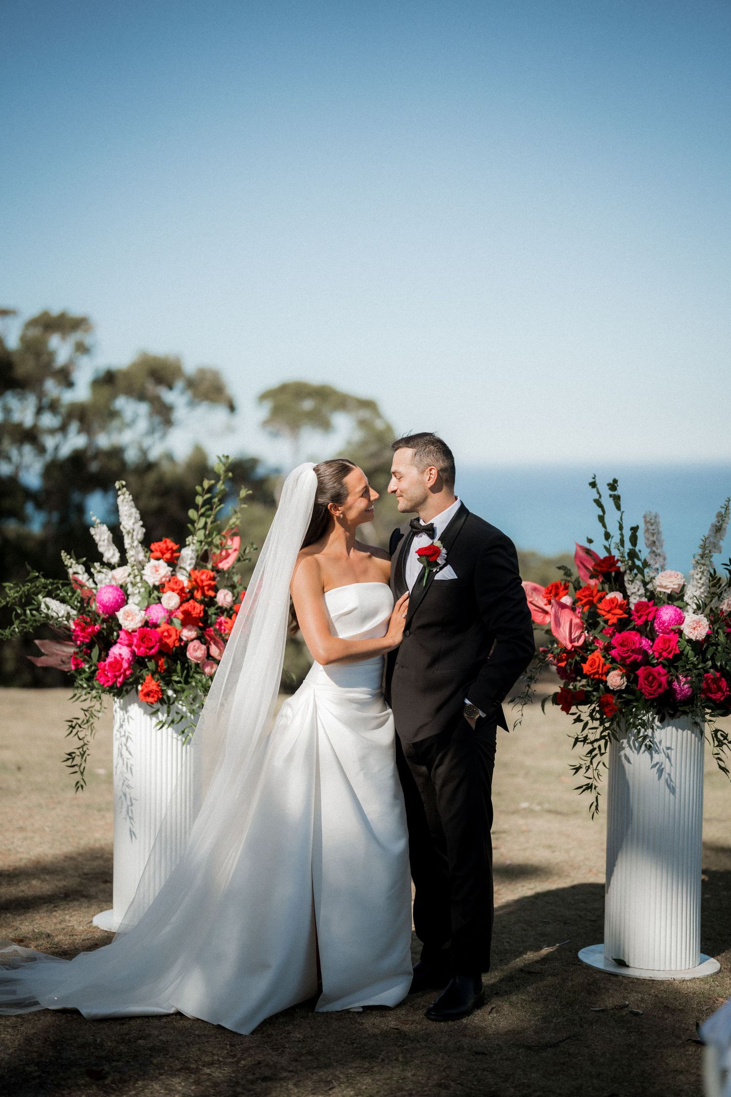 A romantic outdoor wedding ceremony with bright pink floral arrangements on white pillars against an ocean backdrop.