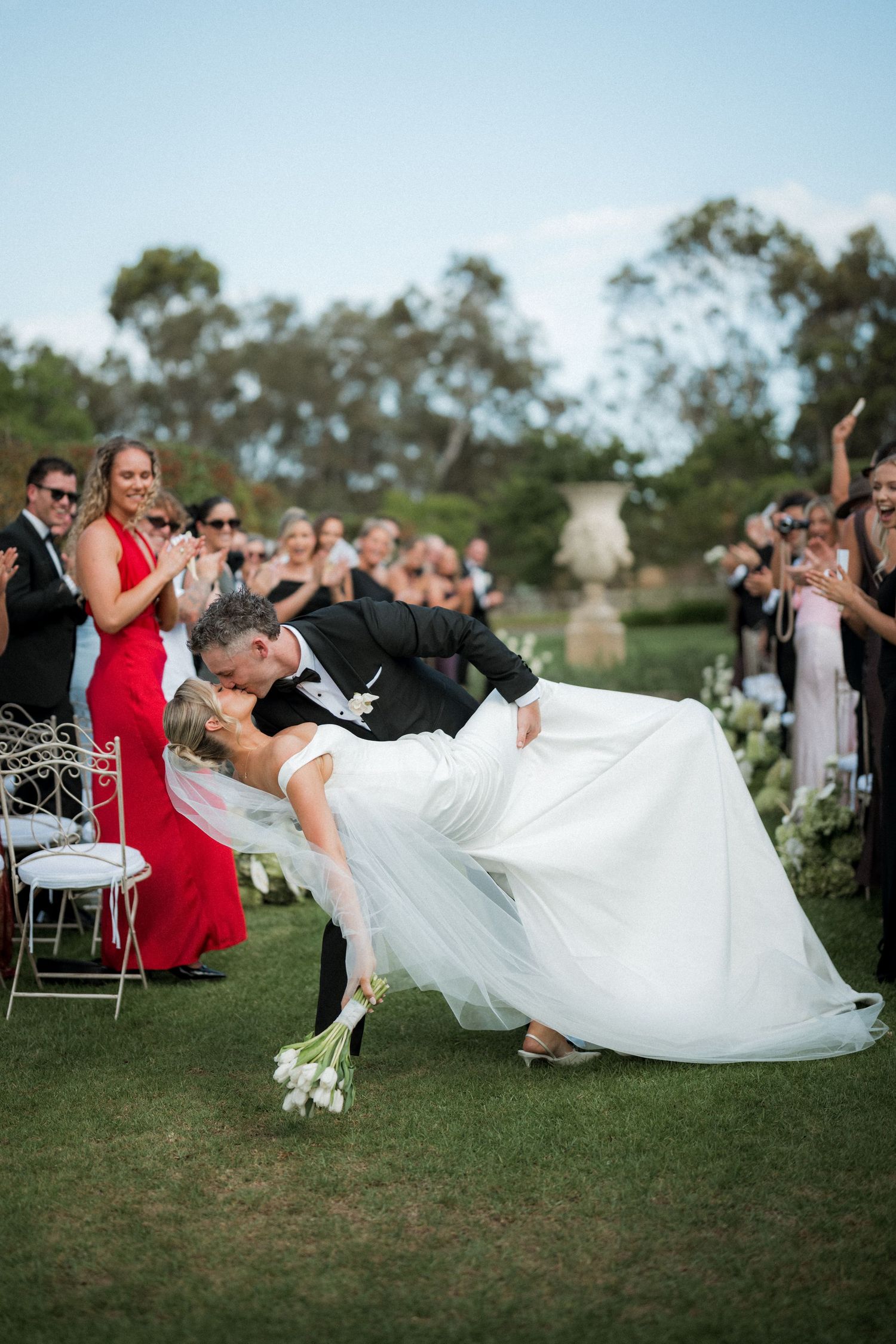Wedding party does synchronized falling pose on grassy lawn during reception.