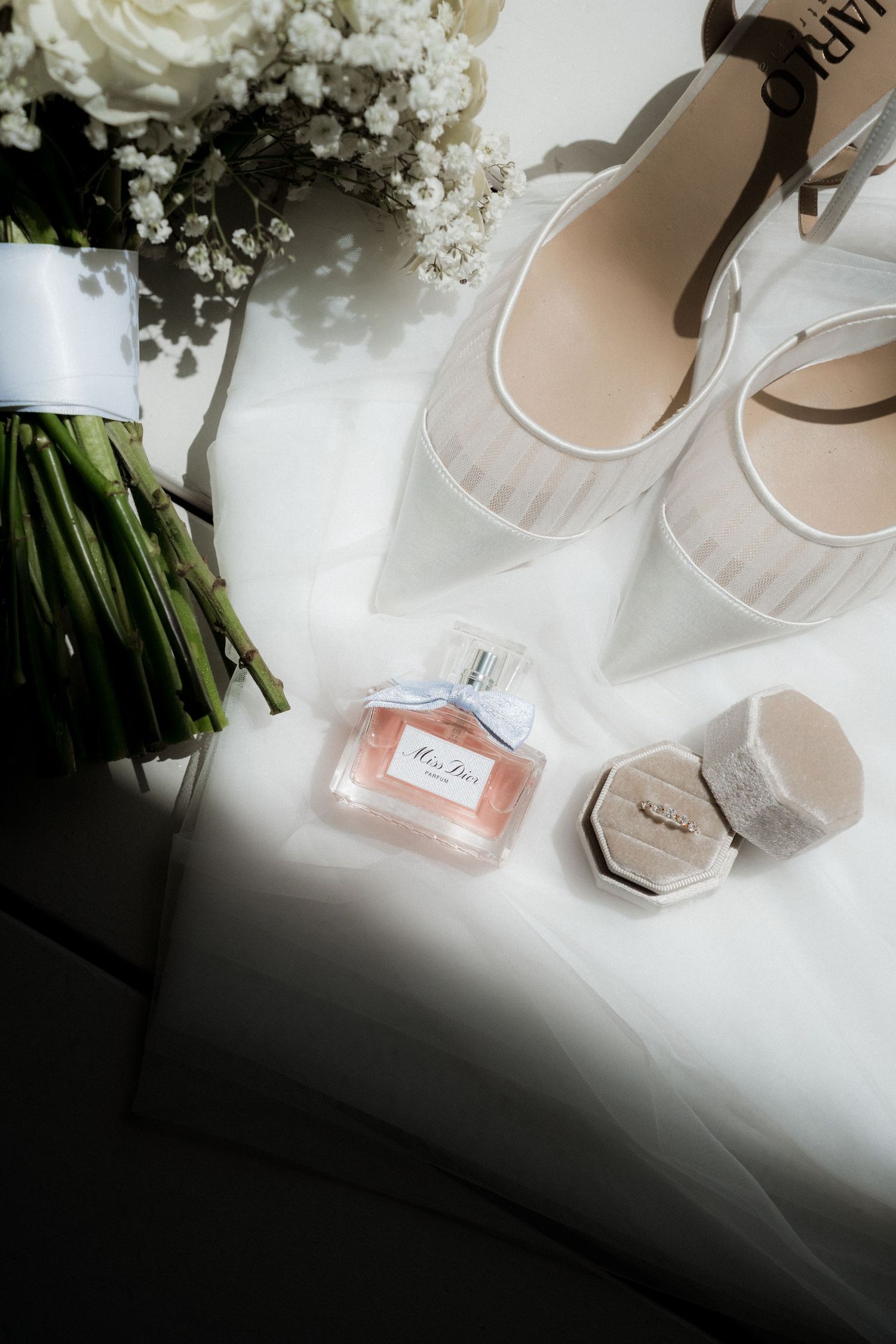 Wedding accessories including white flowers, perfume bottle, and elegant bridal shoes arranged on light surface.