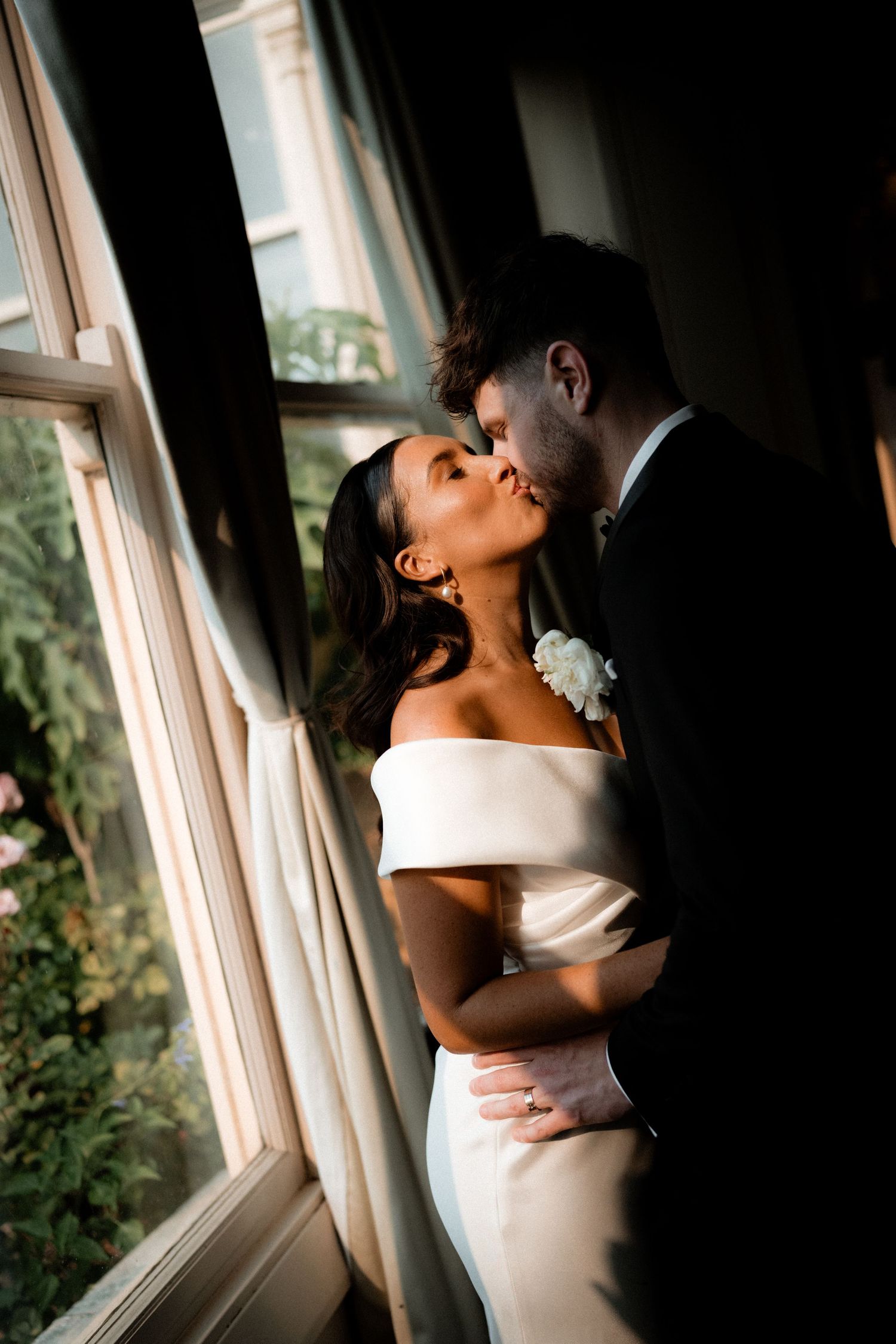 Romantic wedding couple shares intimate moments by sunlit window in elegant venue.