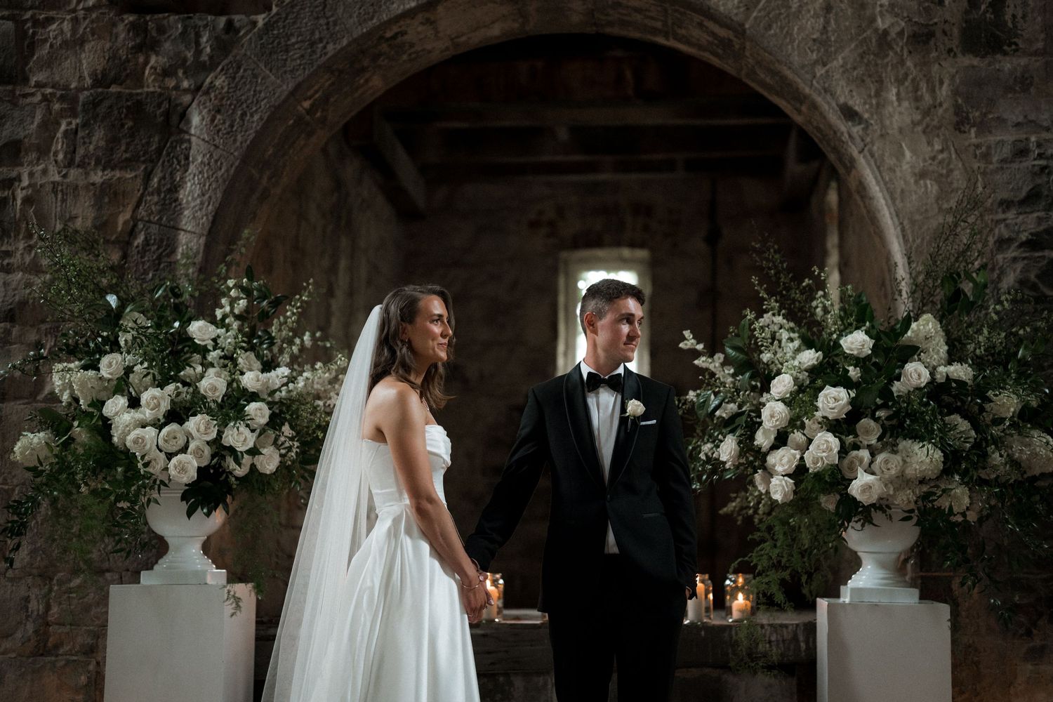 A couple stands beneath a stone archway decorated with elegant white floral arrangements and candles for their wedding ceremony.