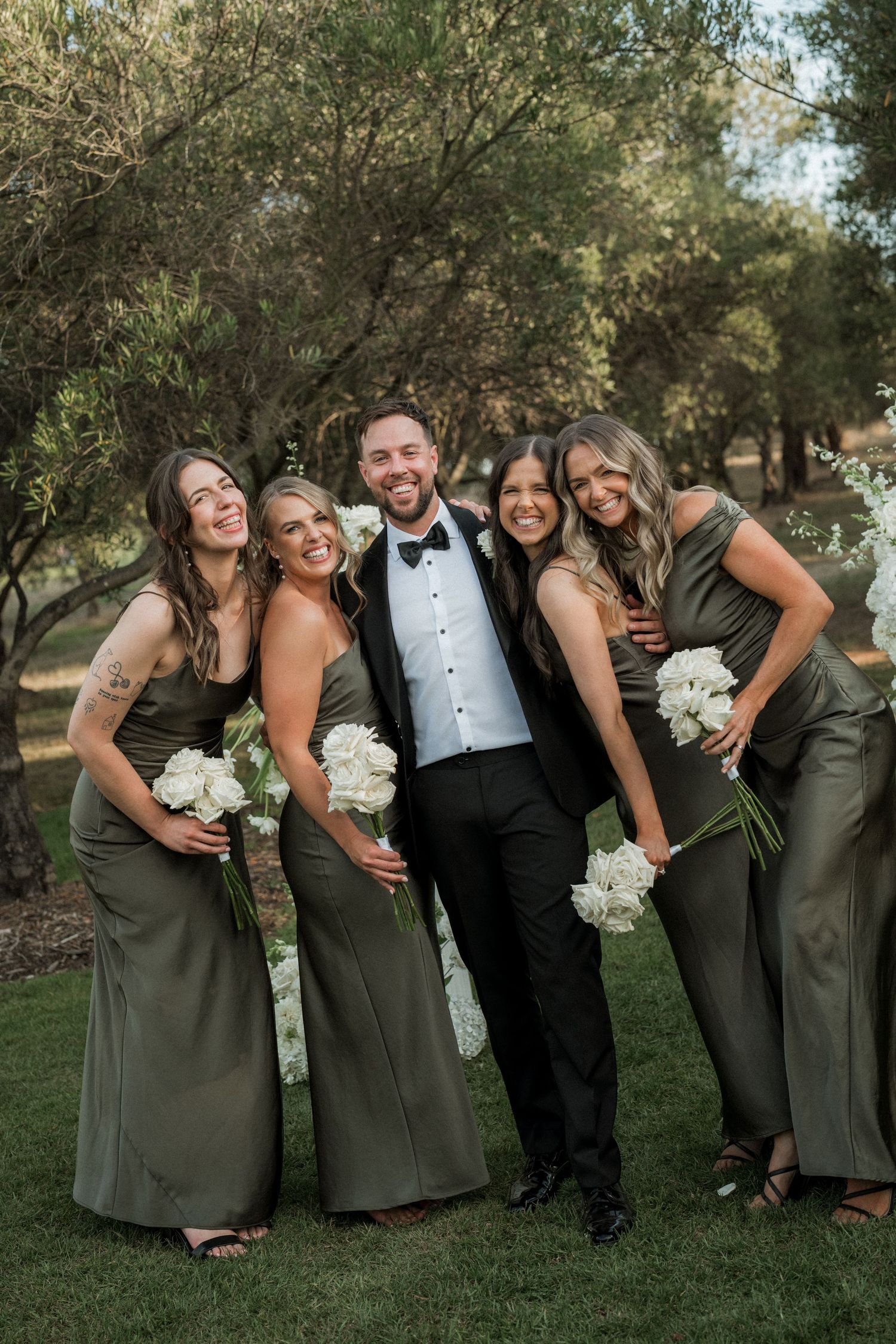 Wedding party in grey dresses and tuxedo pose together on grass with white bouquets.