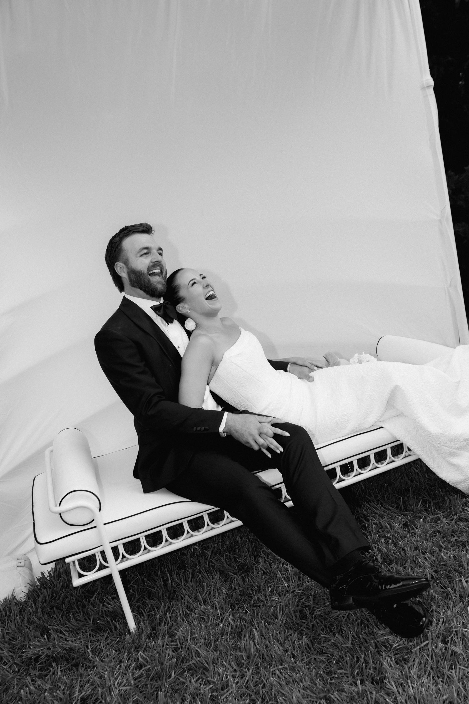 Black and white photo of laughing bride and groom relaxing together on white outdoor bench.