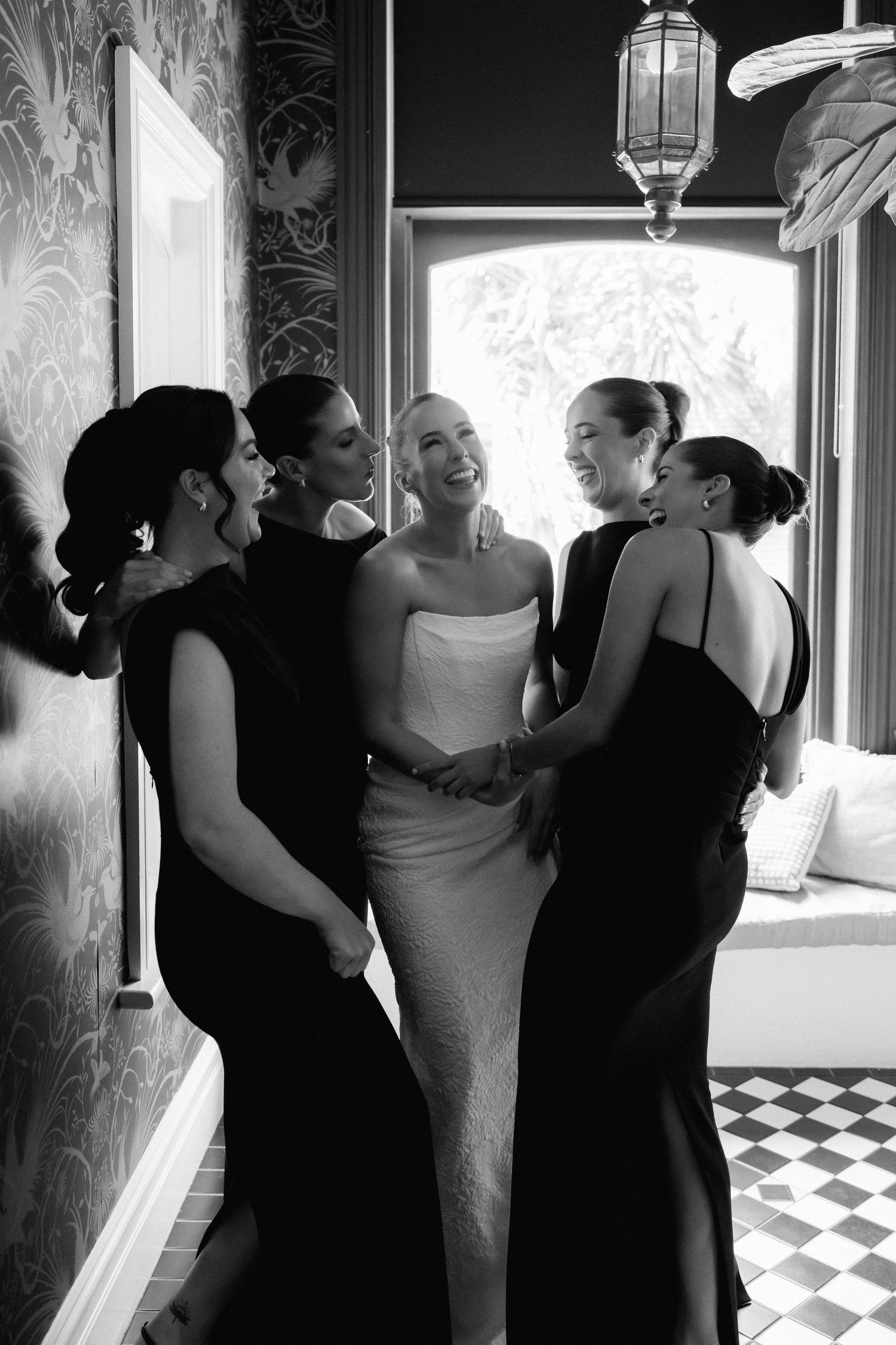 Black and white photo of a bride in white gown surrounded by bridesmaids in black dresses laughing together indoors.