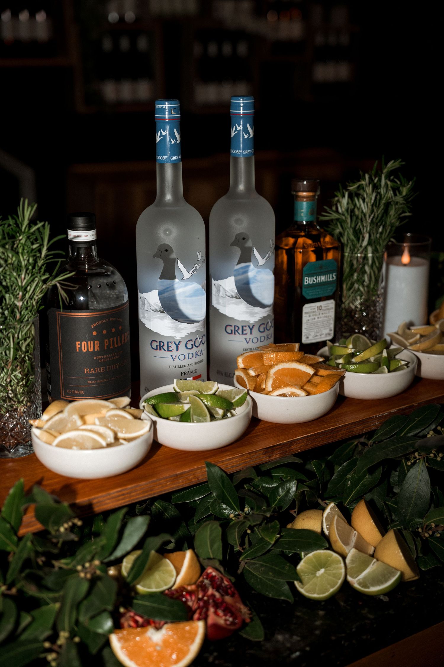 A bar setup with Grey Goose vodka bottles, garnishes, and citrus fruits arranged on a wooden board with fresh herbs.