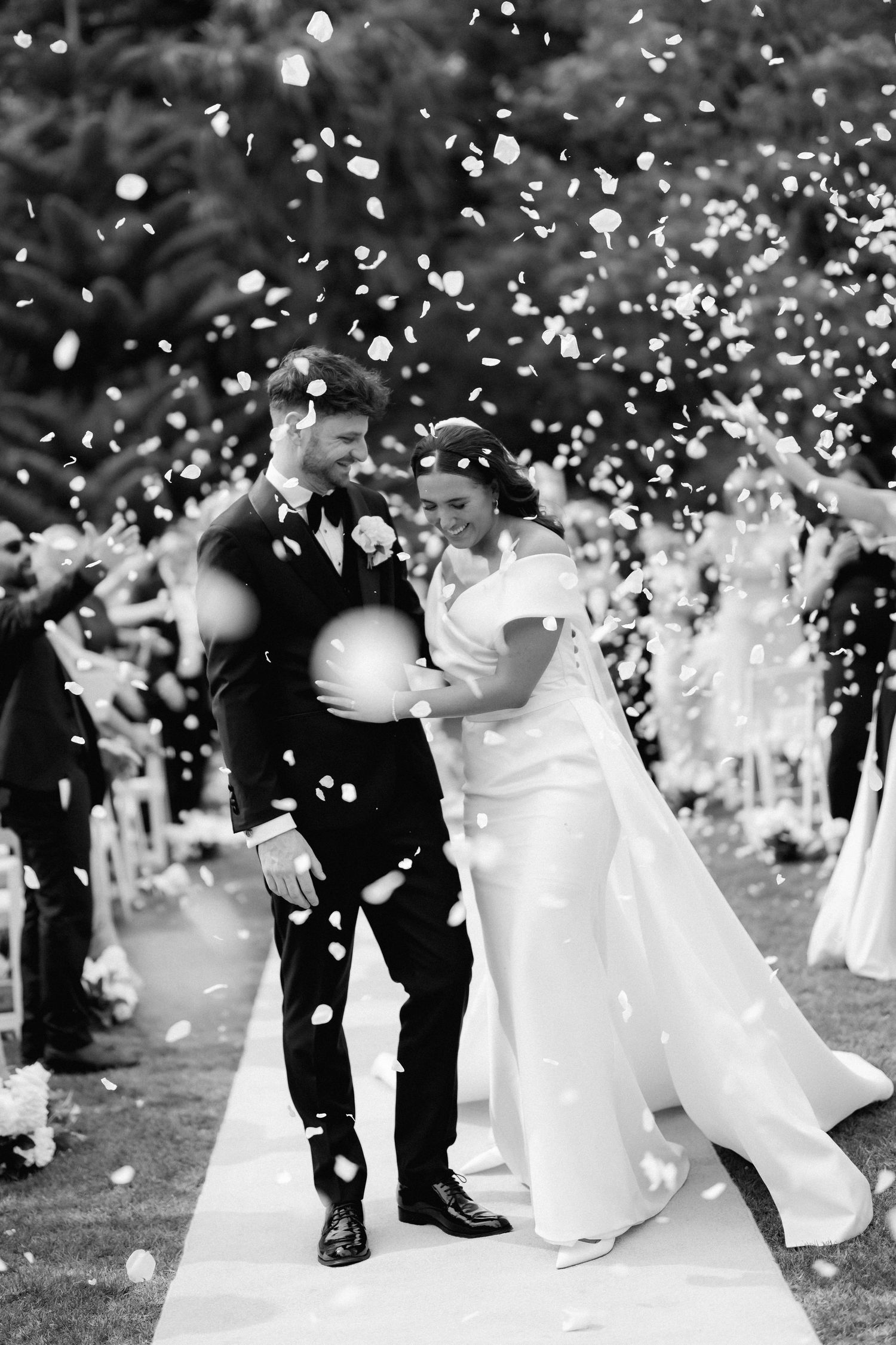 A black and white wedding photo of a couple standing together while confetti falls around them during an outdoor ceremony.