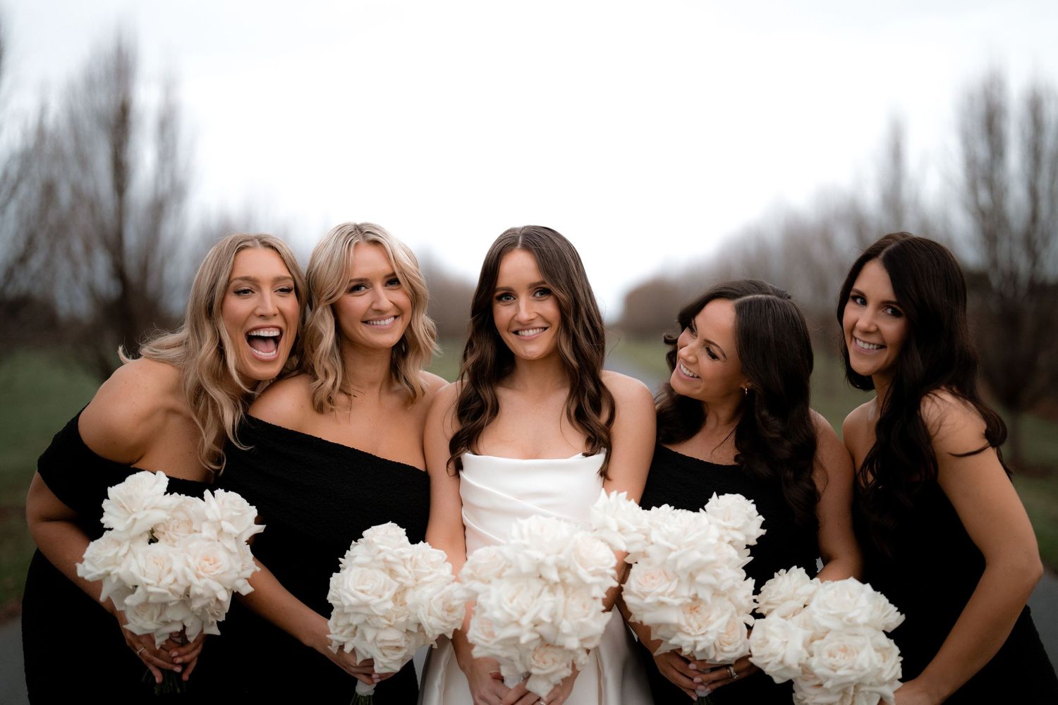 Bridesmaids in black dresses hold white bouquets and laugh together outdoors.