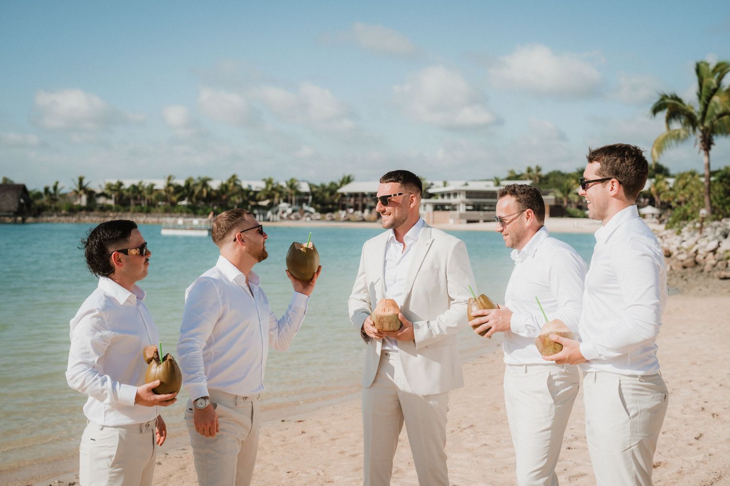Wedding party in all-white attire gather on tropical beach with coconut drinks.