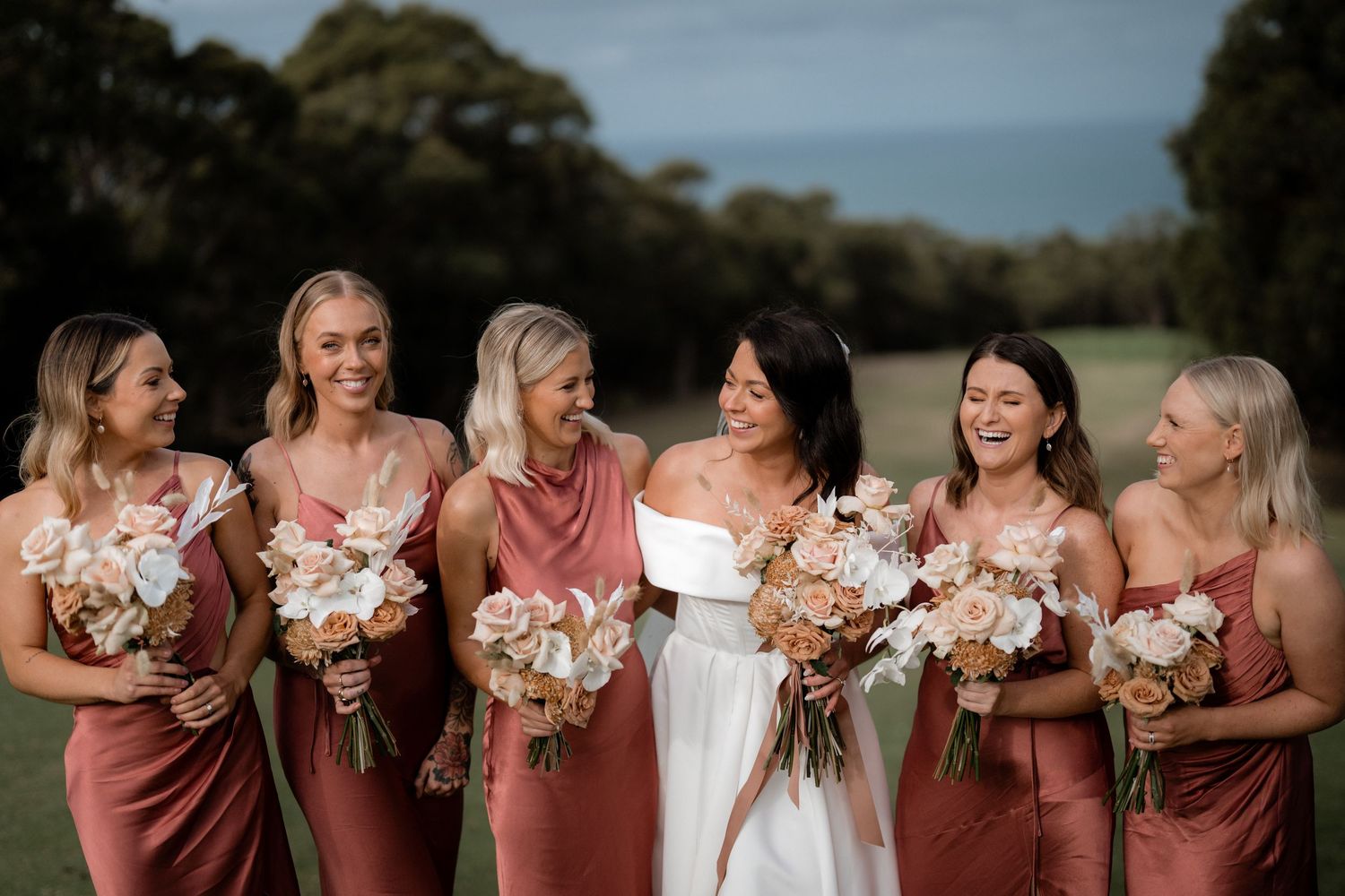 Bridesmaids wearing dusty rose dresses laugh together while holding pastel floral bouquets outdoors at a wedding.
