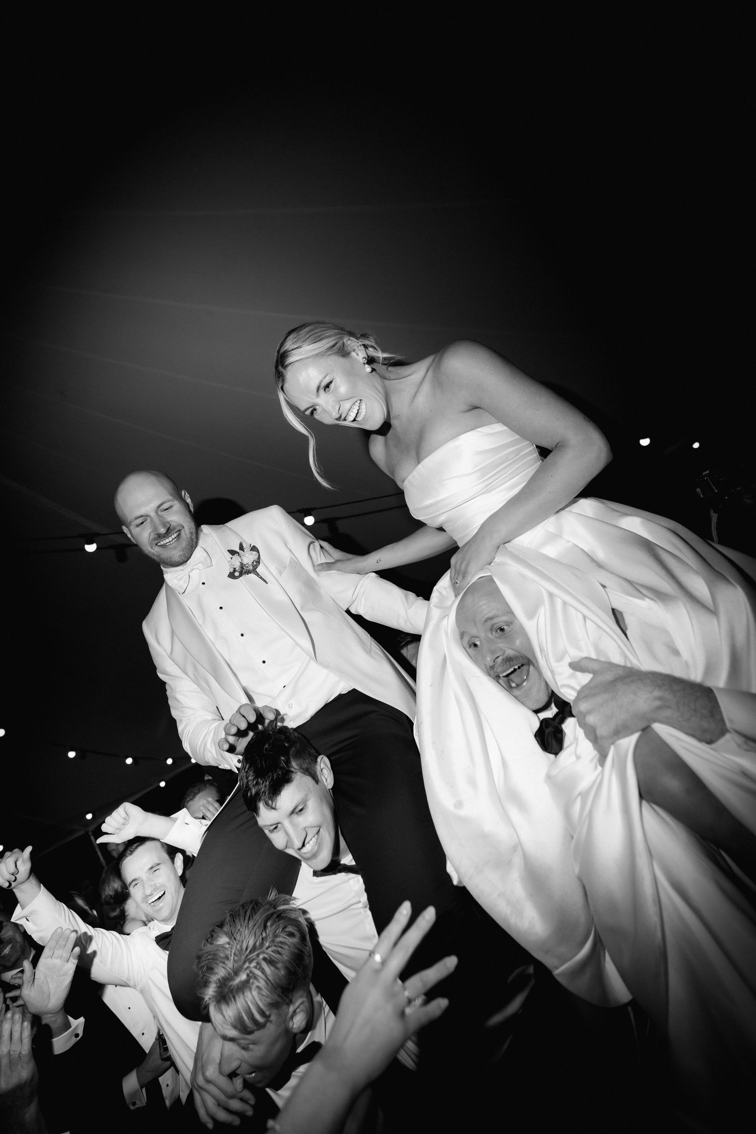 Black and white photo of newlyweds being lifted by celebrating wedding guests during reception.