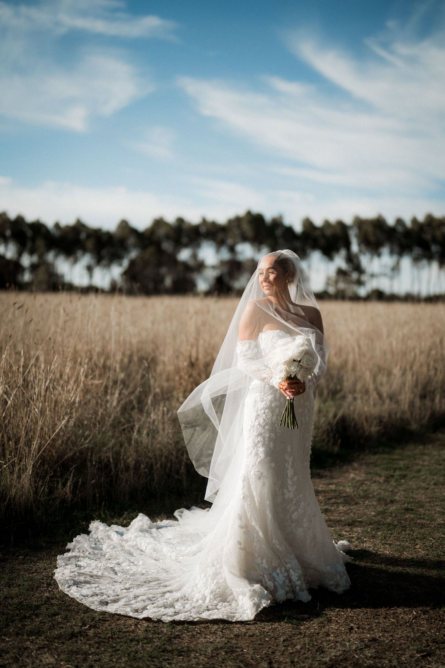 A bride in a flowing white lace wedding dress poses gracefully in a golden field at sunset with palm trees in the distance.