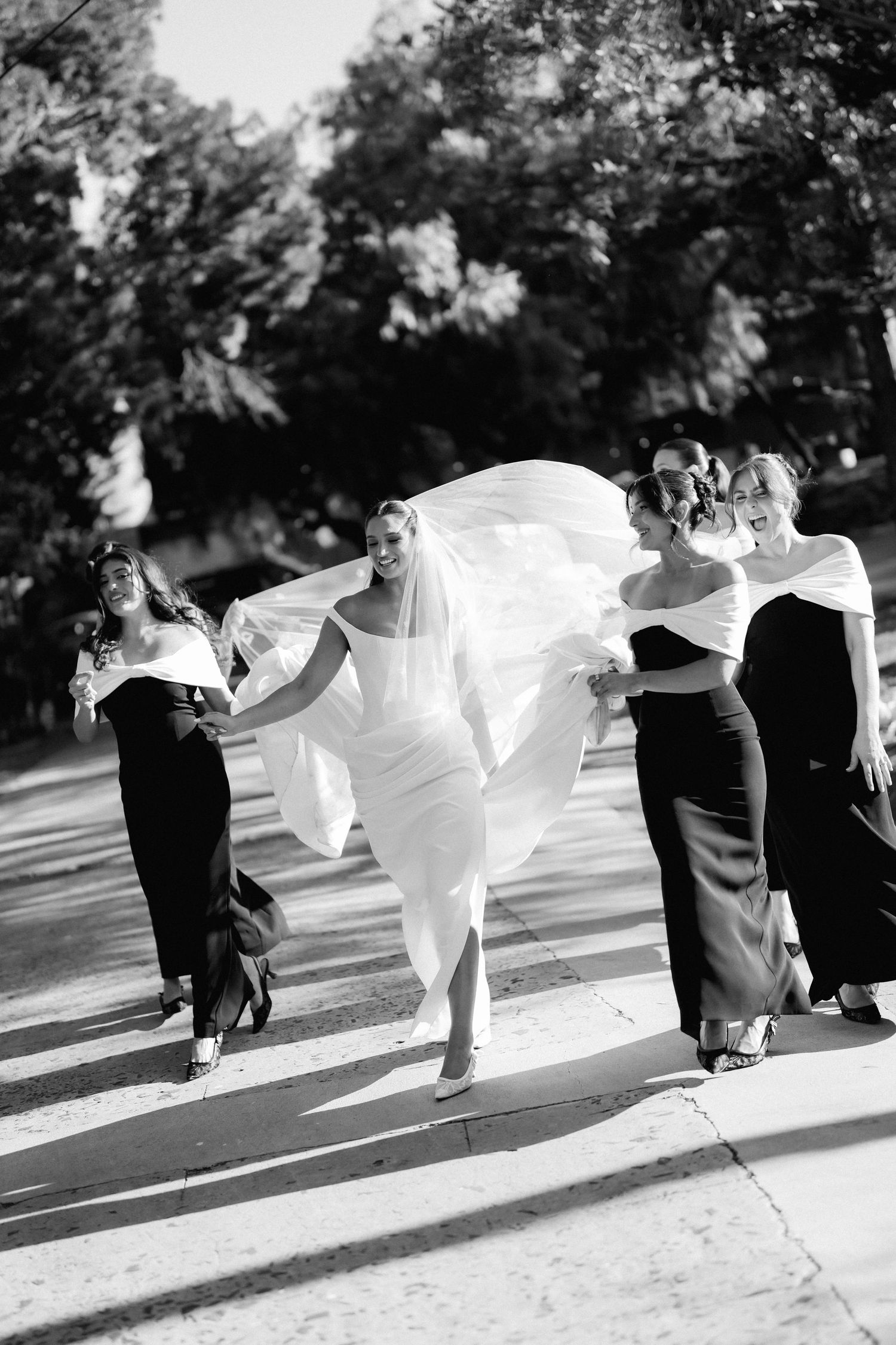 A bride in a flowing white dress runs playfully across a street with bridesmaids in black dresses.