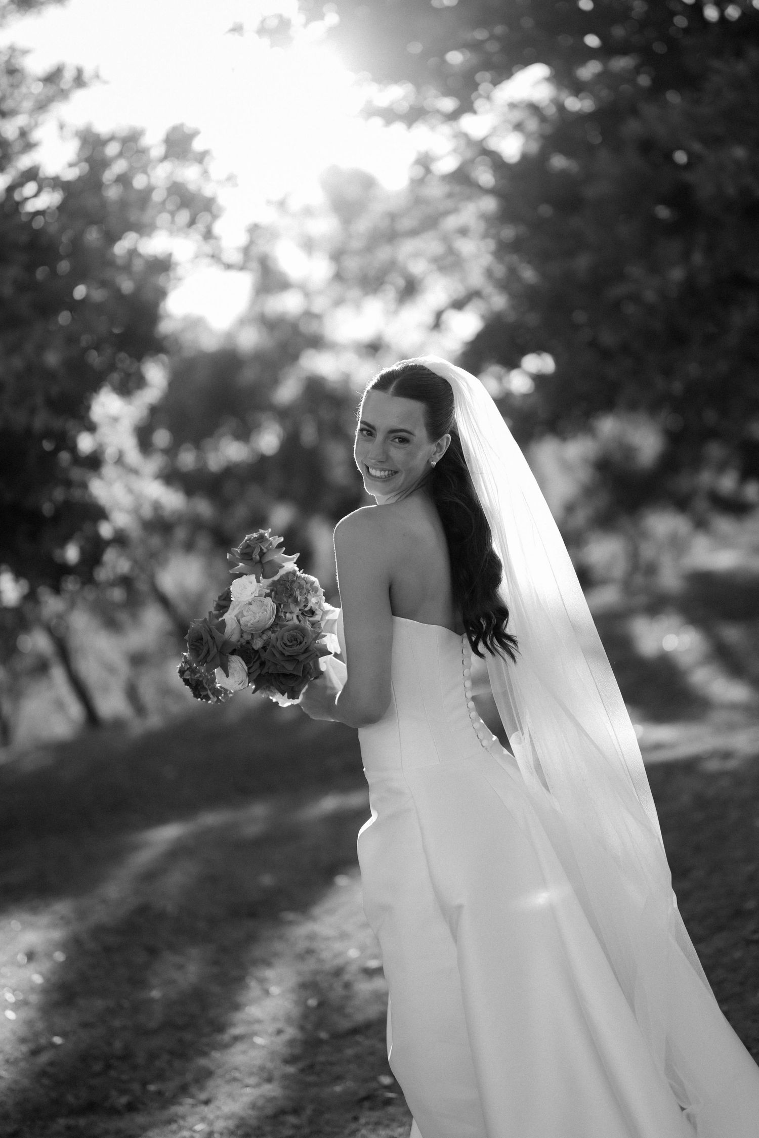 A bride in a white wedding dress holds a bouquet while posing outdoors in black and white photography.