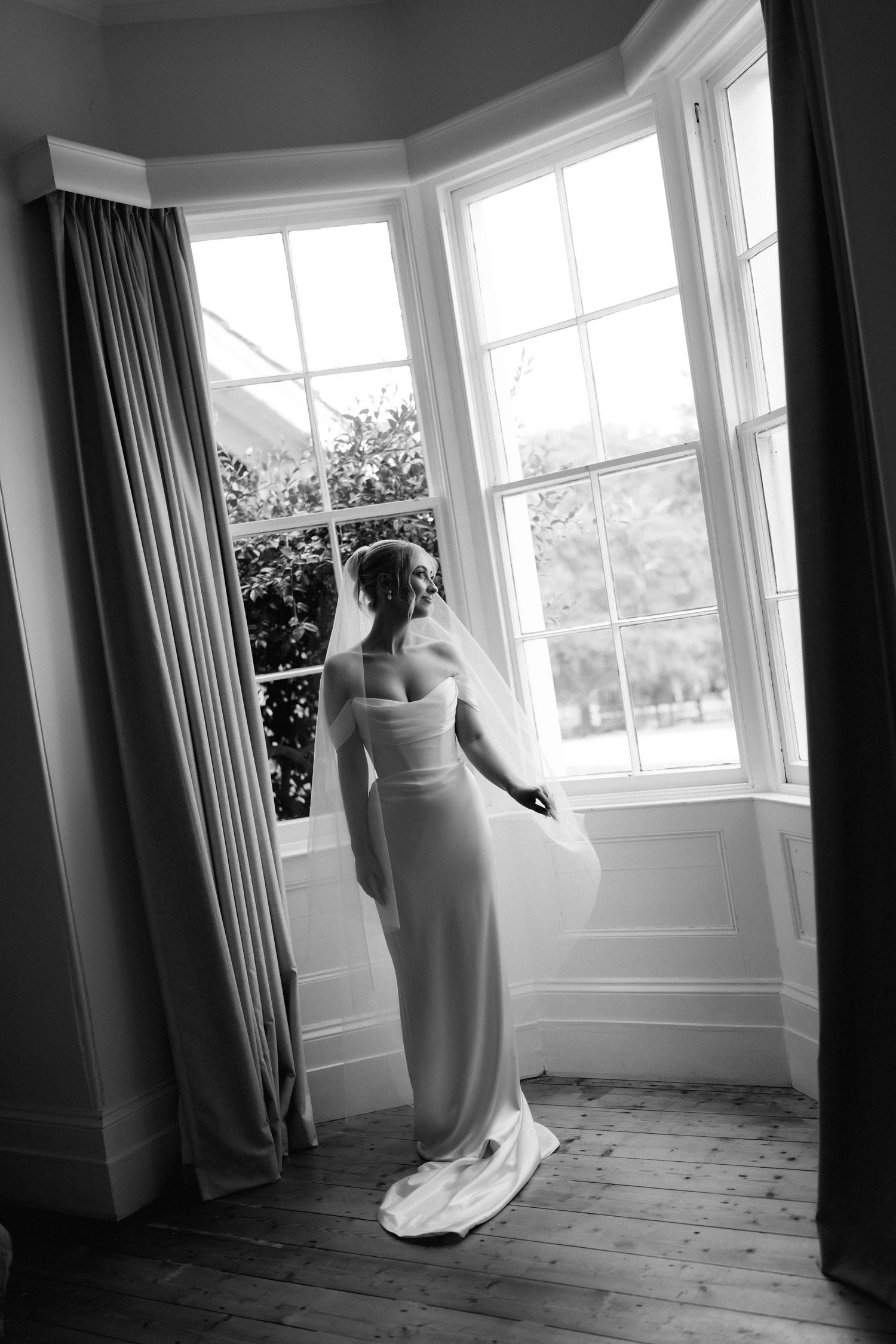 A black and white photo of an elegant bride in a sleek gown standing by a bay window with flowing curtains and wooden floors.