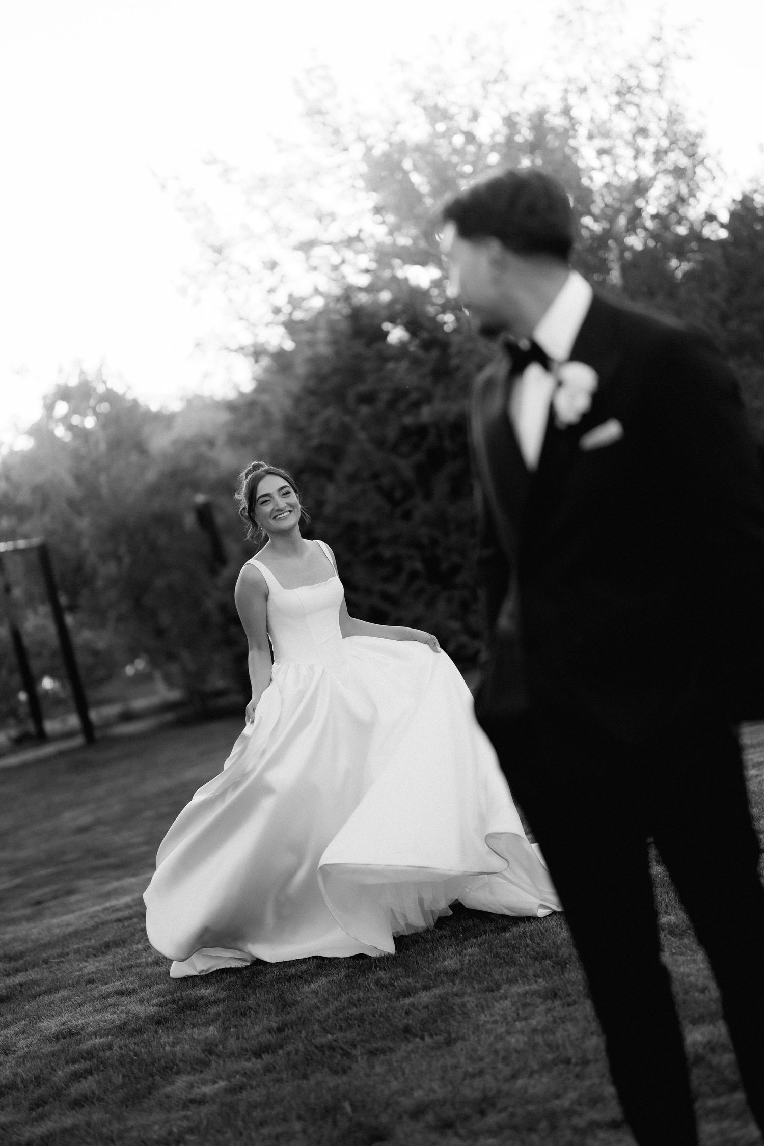 Black and white photo of a smiling bride in a satin gown twirling on a lawn as the groom in a tuxedo looks on.