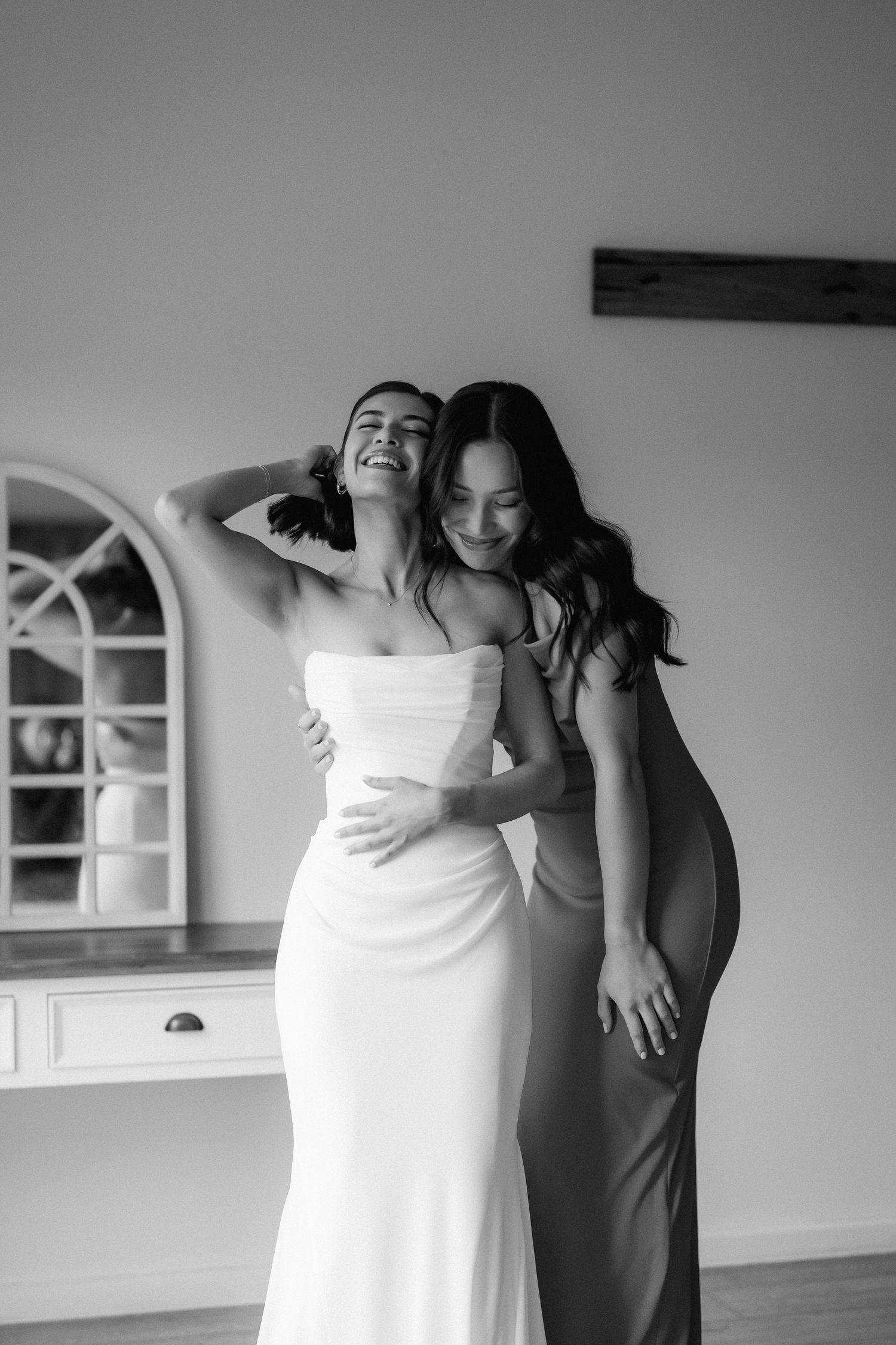 Black and white photo of a bride in a white gown laughing as her bridesmaid hugs her from behind.