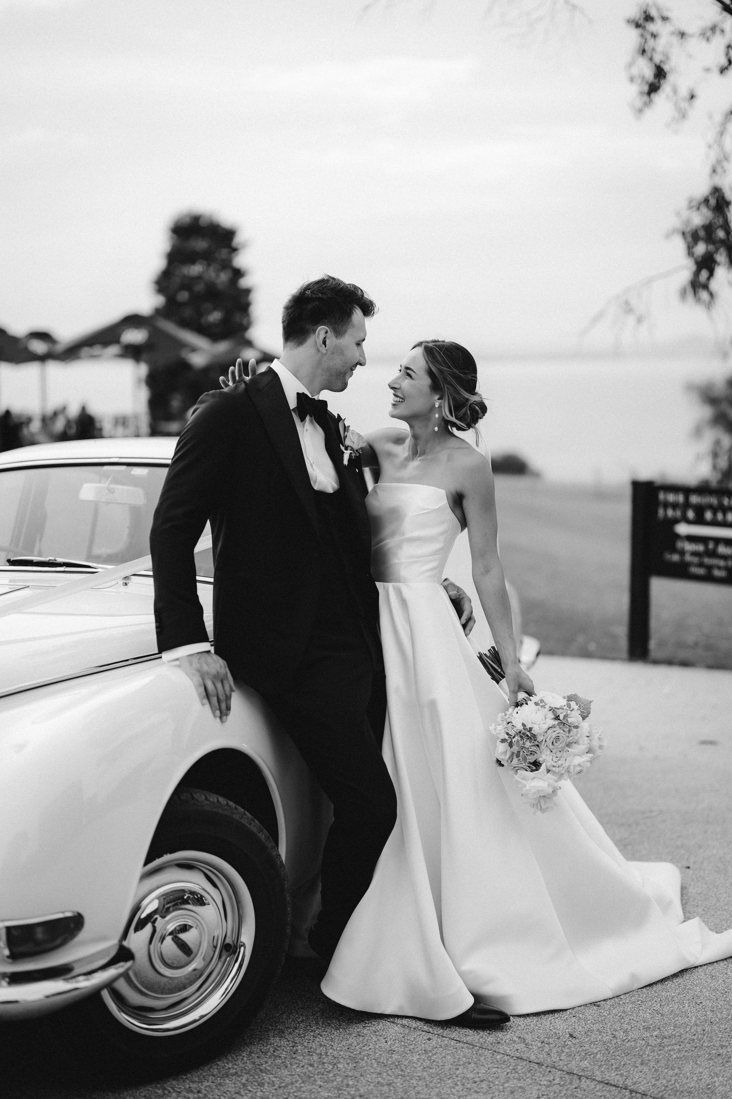 A black and white wedding photo with a couple posing beside a vintage car, holding a floral bouquet.