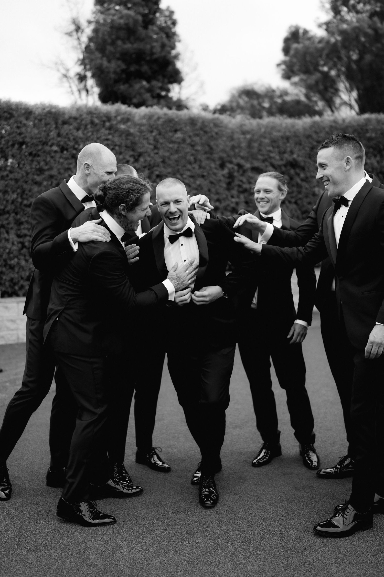 Black and white photo of groomsmen in tuxedos goofing around and laughing together outdoors.