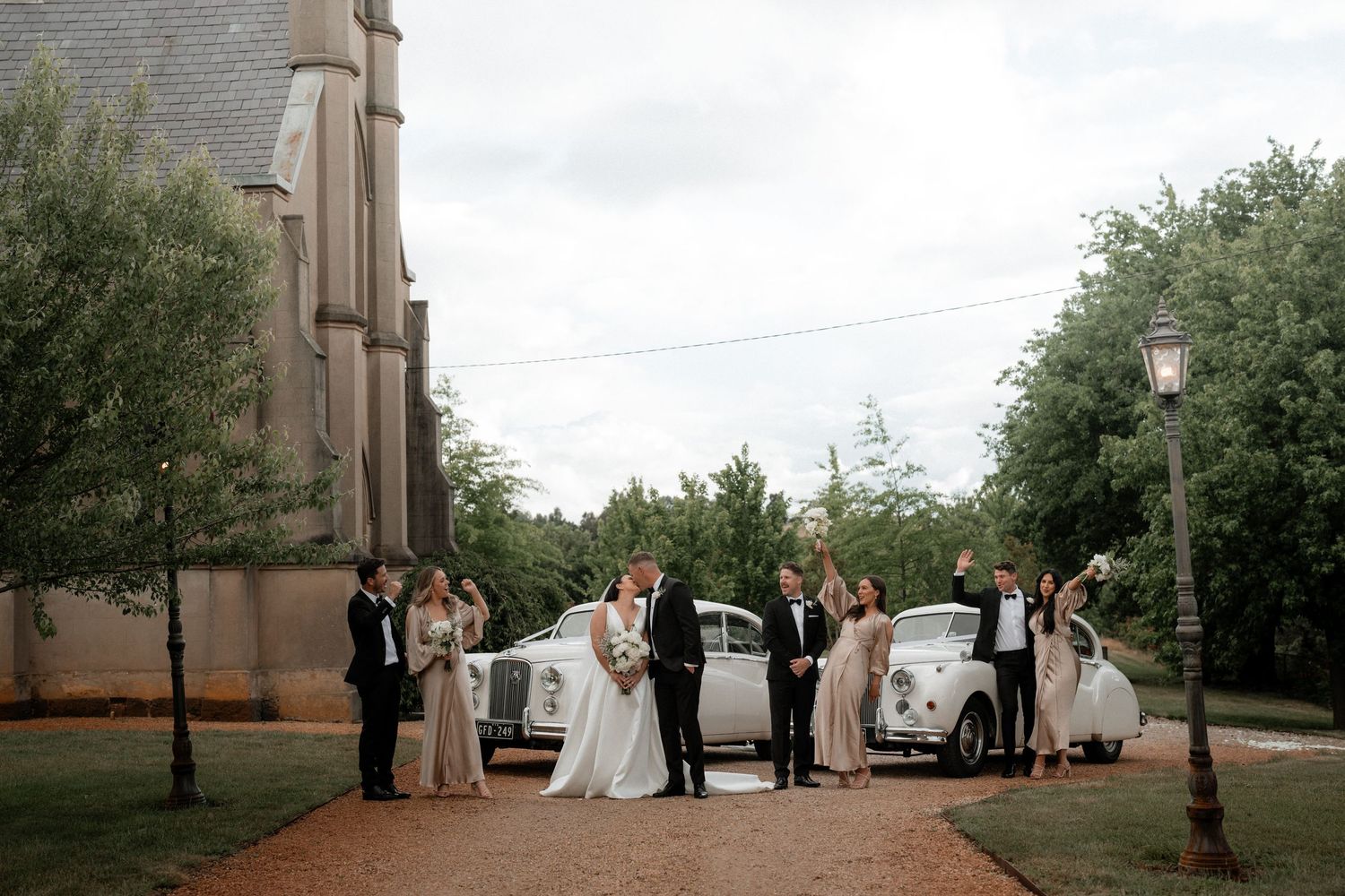 A wedding party poses on a tree-lined path beside two vintage white cars outside a stone church.