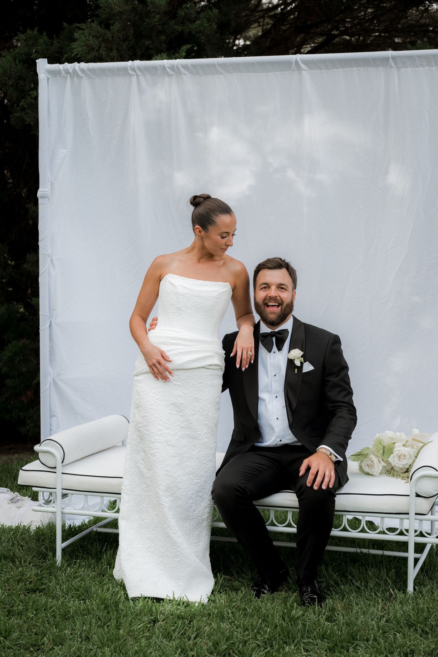 Smiling bride and groom posing on white outdoor bench against white backdrop on grass.