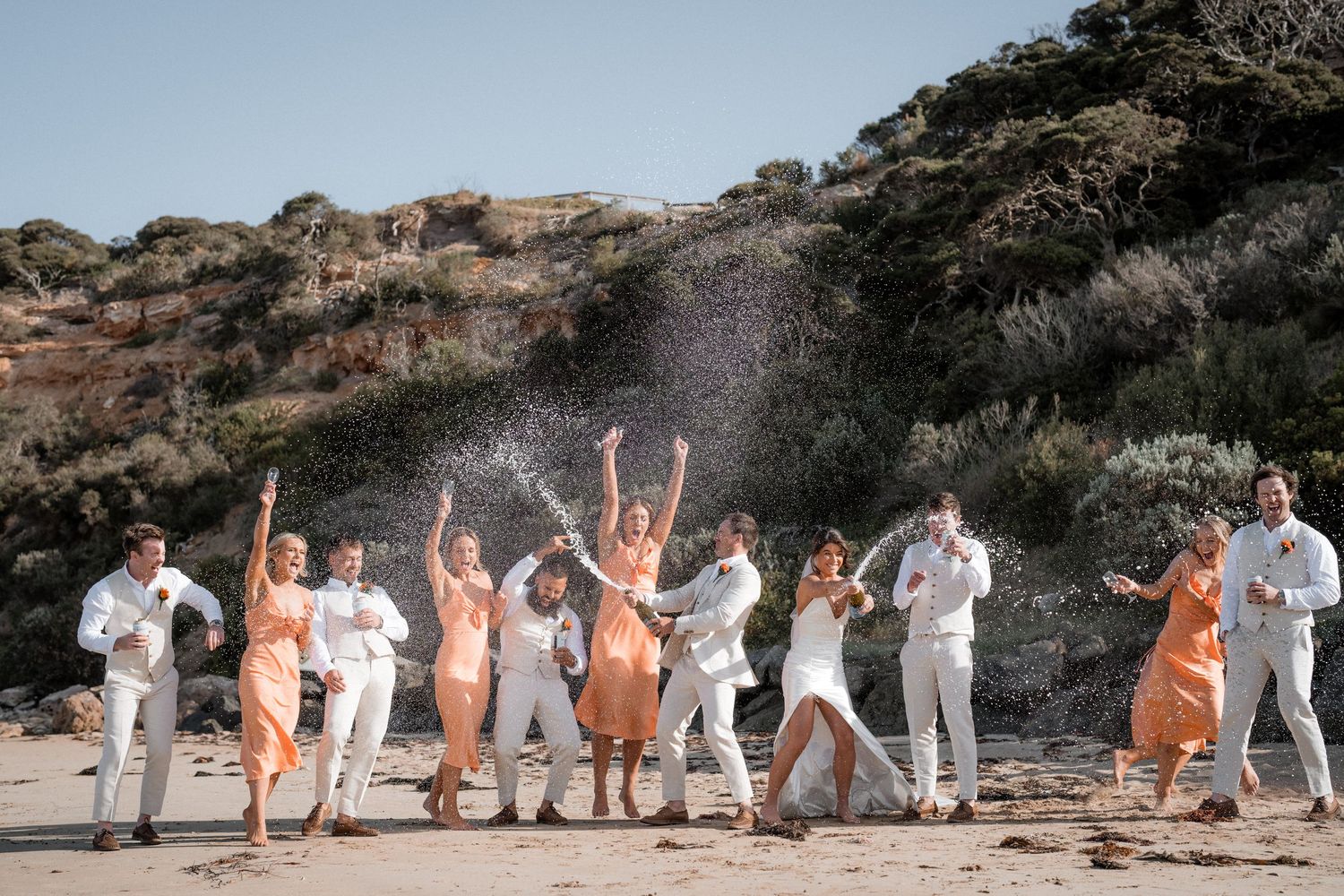 Wedding party posing on rocky beach in white and peach attire.